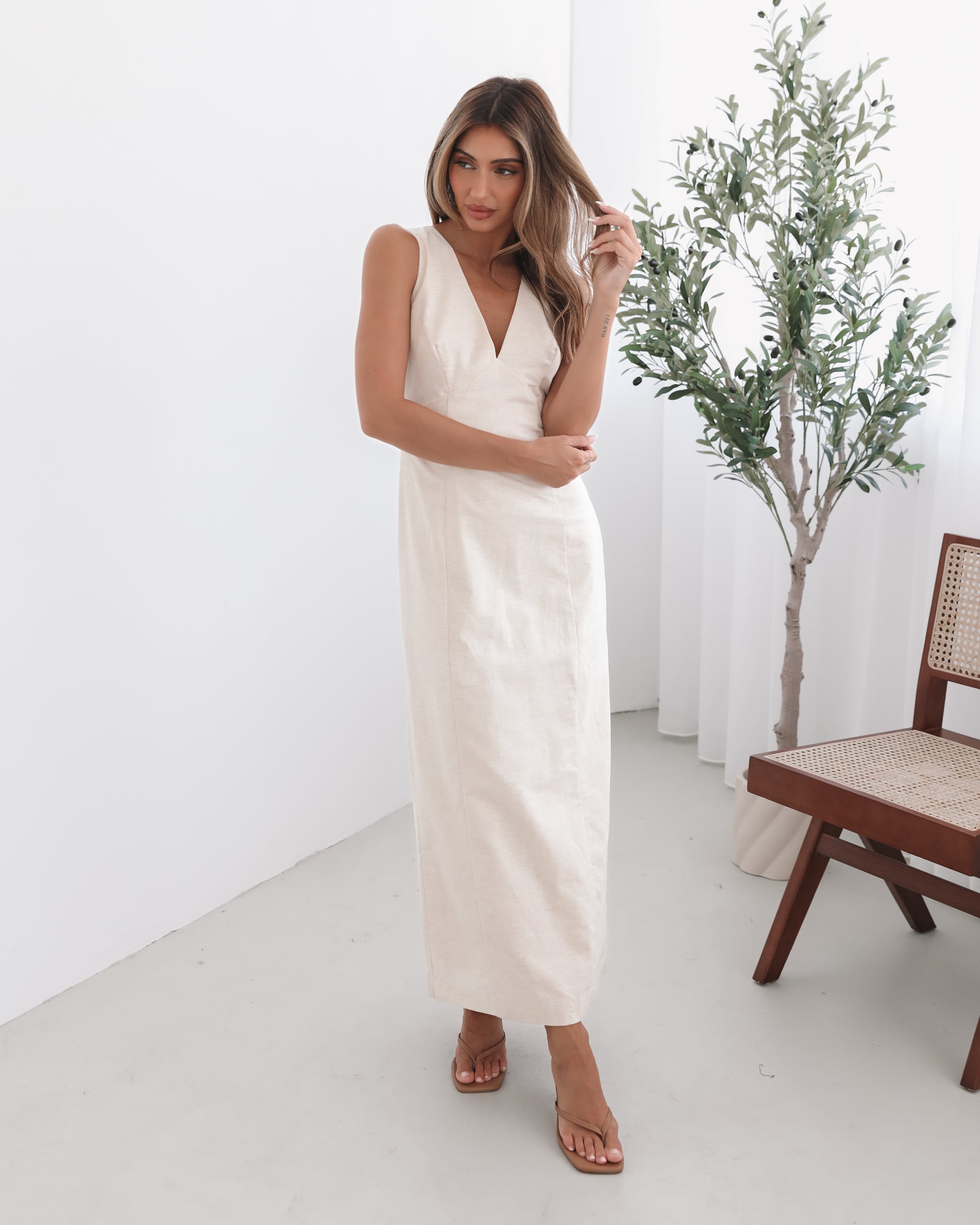 A woman wearing a beige maxi dress with a plunge neckline, bow and slit at the back in a white setting with a wooden chair and a plant