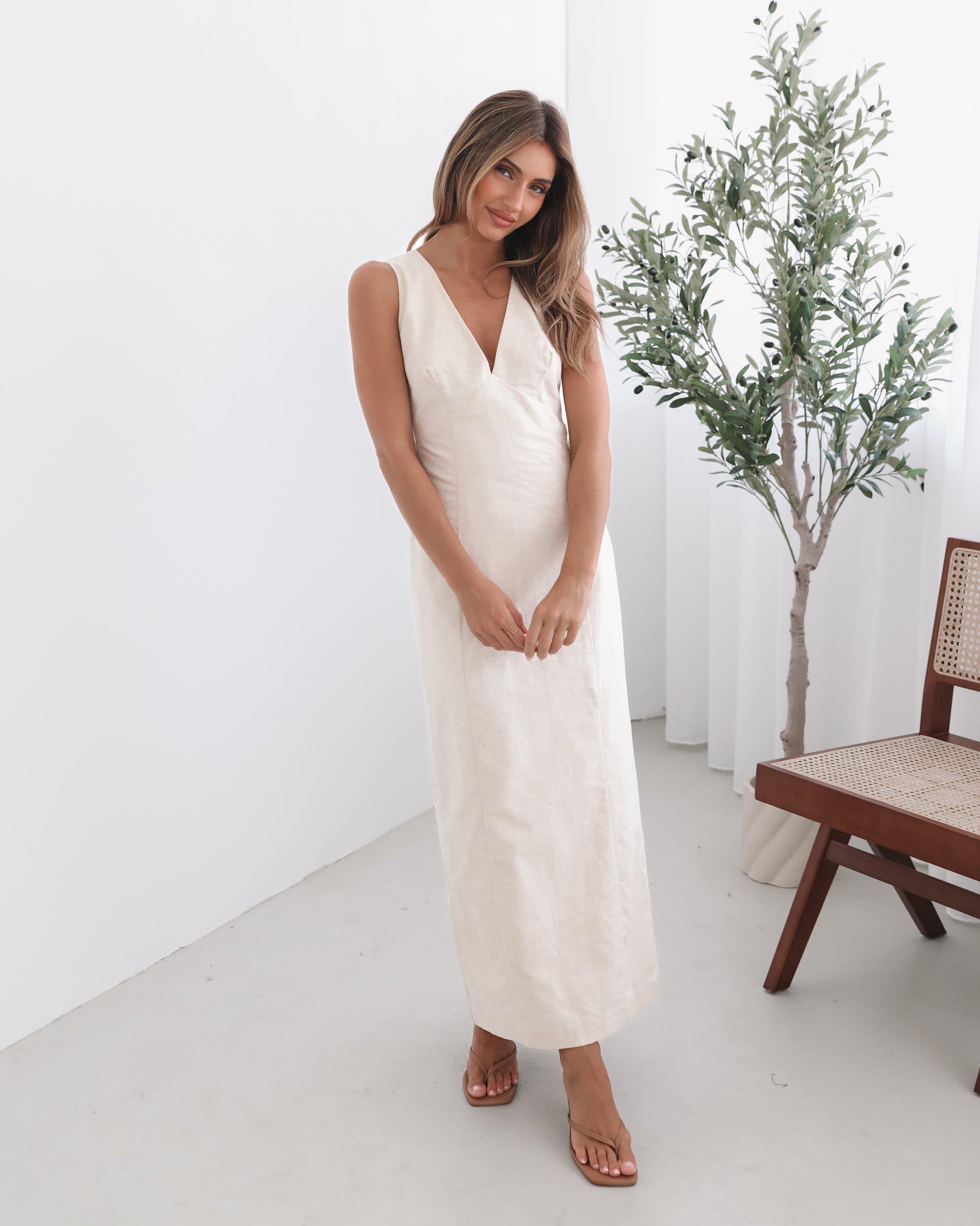 A woman wearing a beige maxi dress with a plunge neckline, bow and slit at the back in a white setting with a wooden chair and a plant