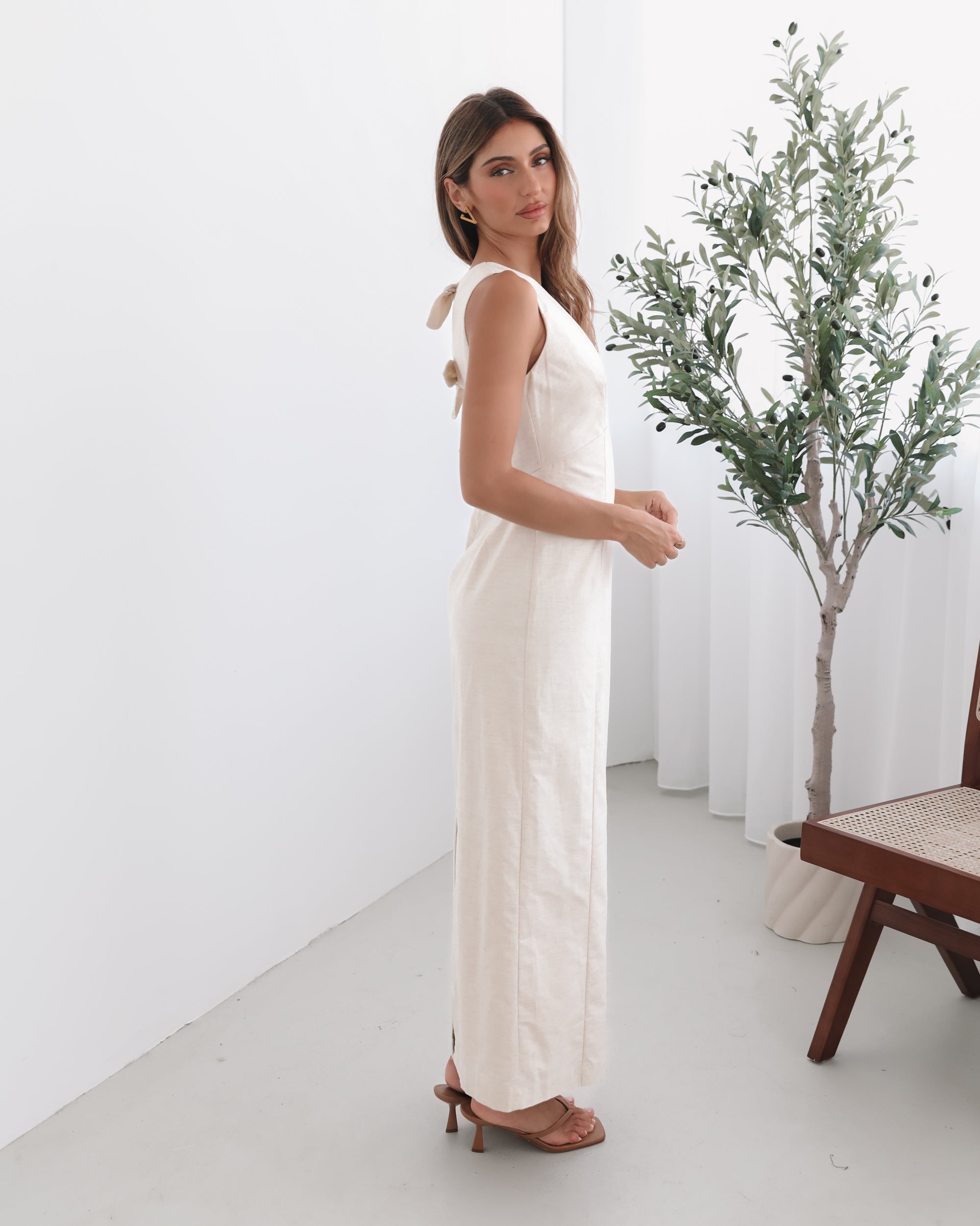 A woman wearing a beige maxi dress with a plunge neckline, bow and slit at the back in a white setting with a wooden chair and a plant