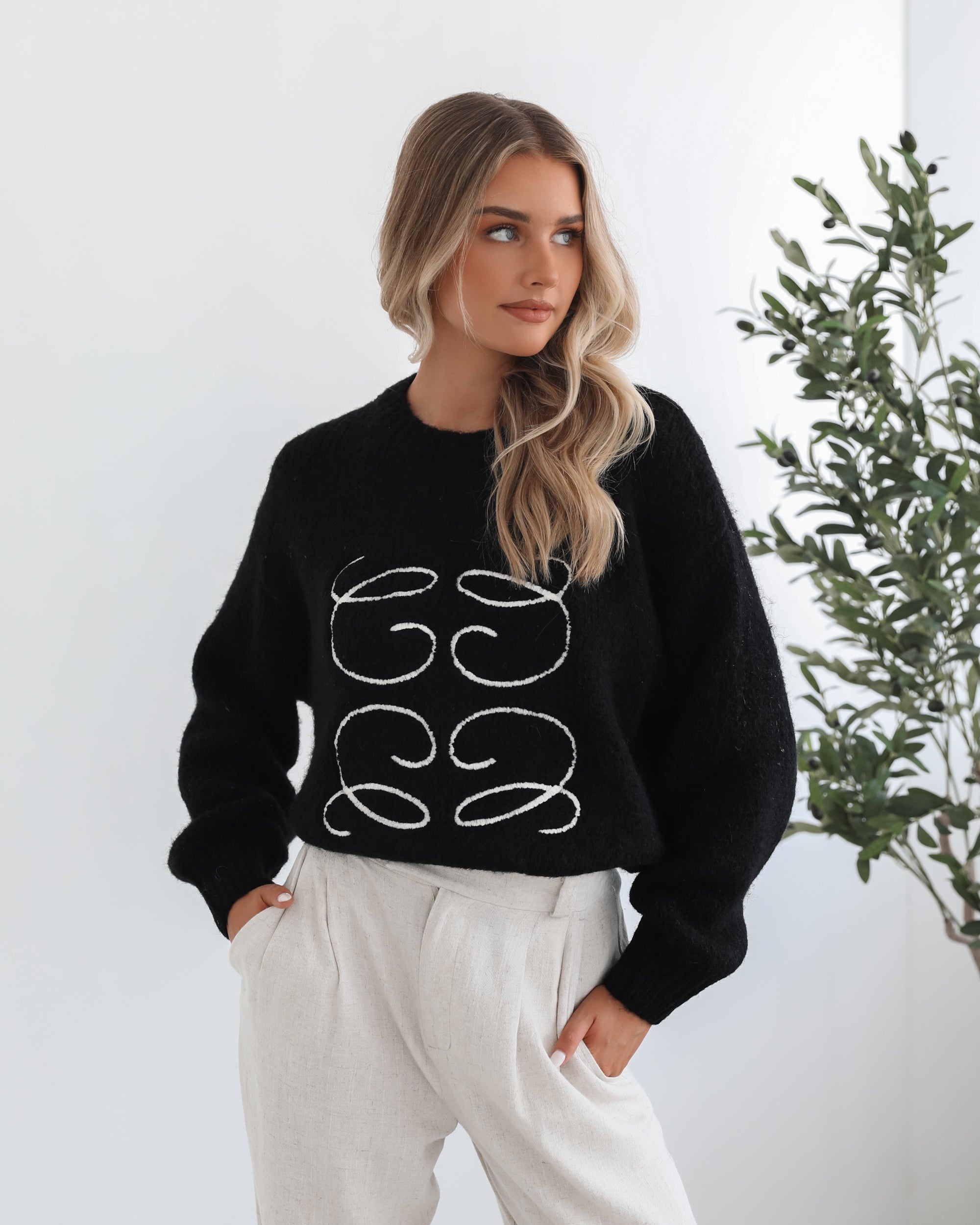 A woman with long, wavy blonde hair is standing indoors near a potted plant, wearing a black sweater with a white abstract design on the front and high-waisted blue jeans. She is looking at the camera with a neutral expression and has one hand resting on her hip. The background is minimalistic with white walls.