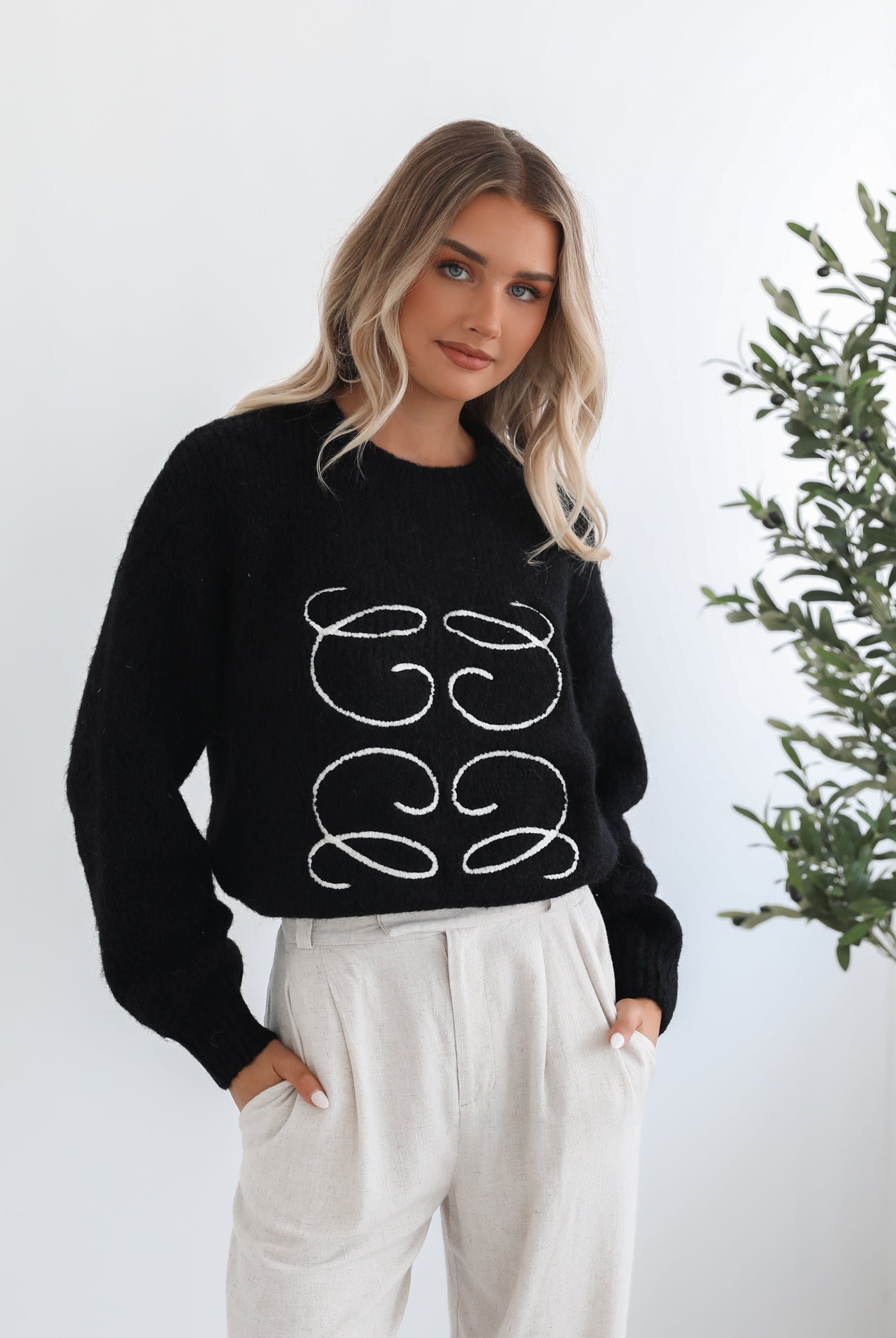 A woman with long, wavy blonde hair is standing indoors near a potted plant, wearing a black sweater with a white abstract design on the front and high-waisted blue jeans. She is looking at the camera with a neutral expression and has one hand resting on her hip. The background is minimalistic with white walls.