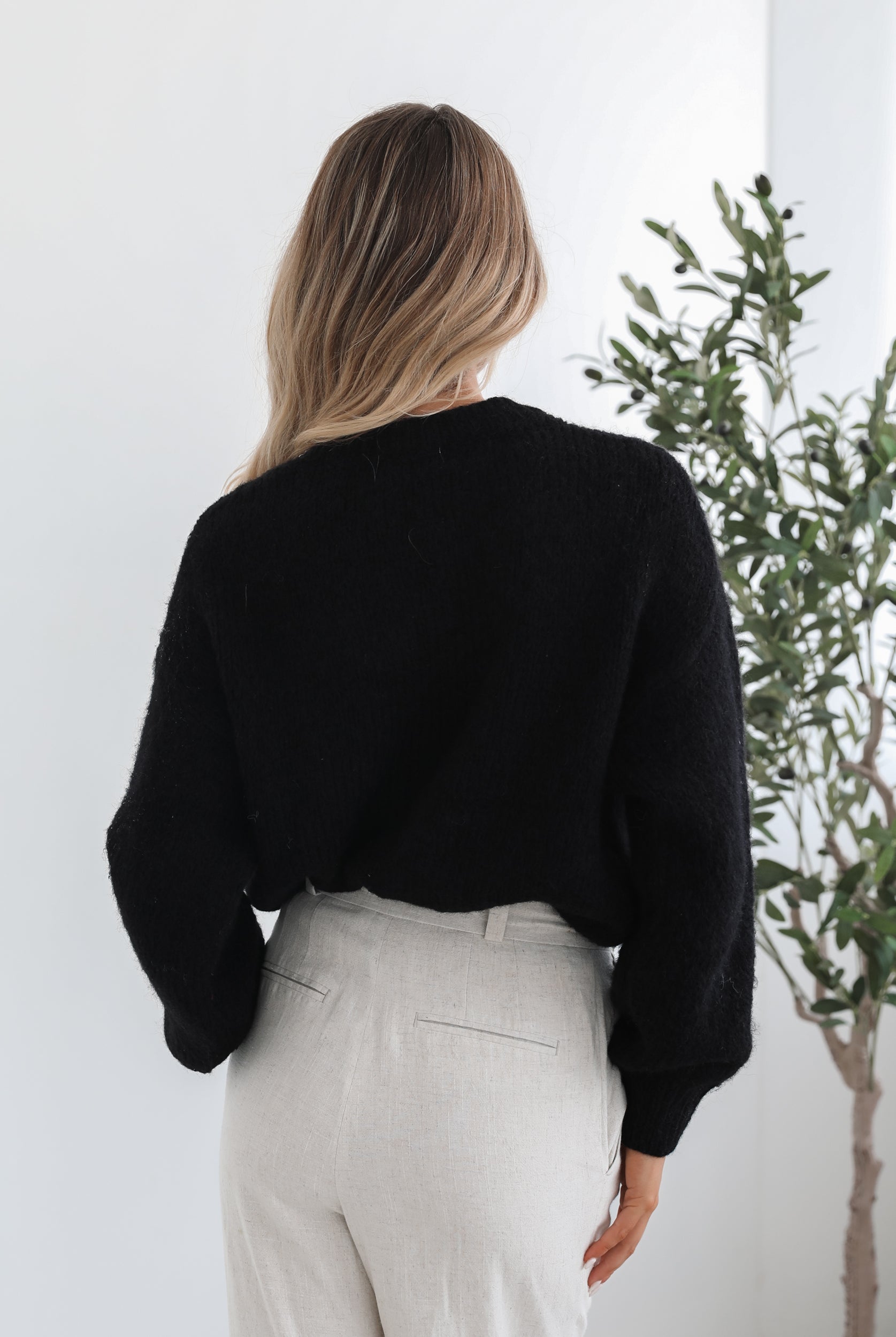 A woman with long, wavy blonde hair is standing indoors near a potted plant, wearing a black sweater with a white abstract design on the front and high-waisted blue jeans. She is looking at the camera with a neutral expression and has one hand resting on her hip. The background is minimalistic with white walls.