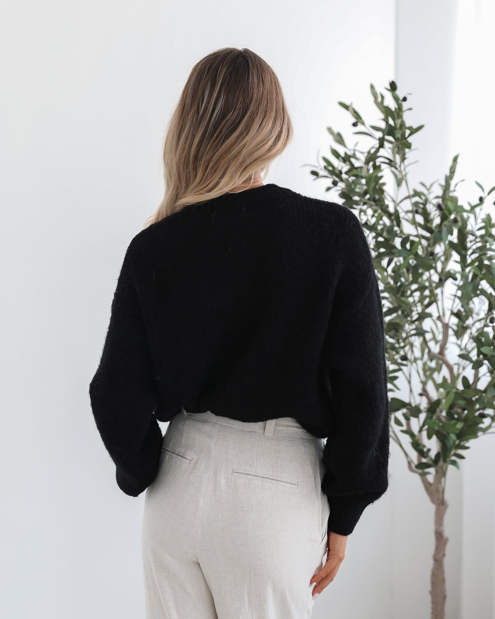 A woman with long, wavy blonde hair is standing indoors near a potted plant, wearing a black sweater with a white abstract design on the front and high-waisted blue jeans. She is looking at the camera with a neutral expression and has one hand resting on her hip. The background is minimalistic with white walls.