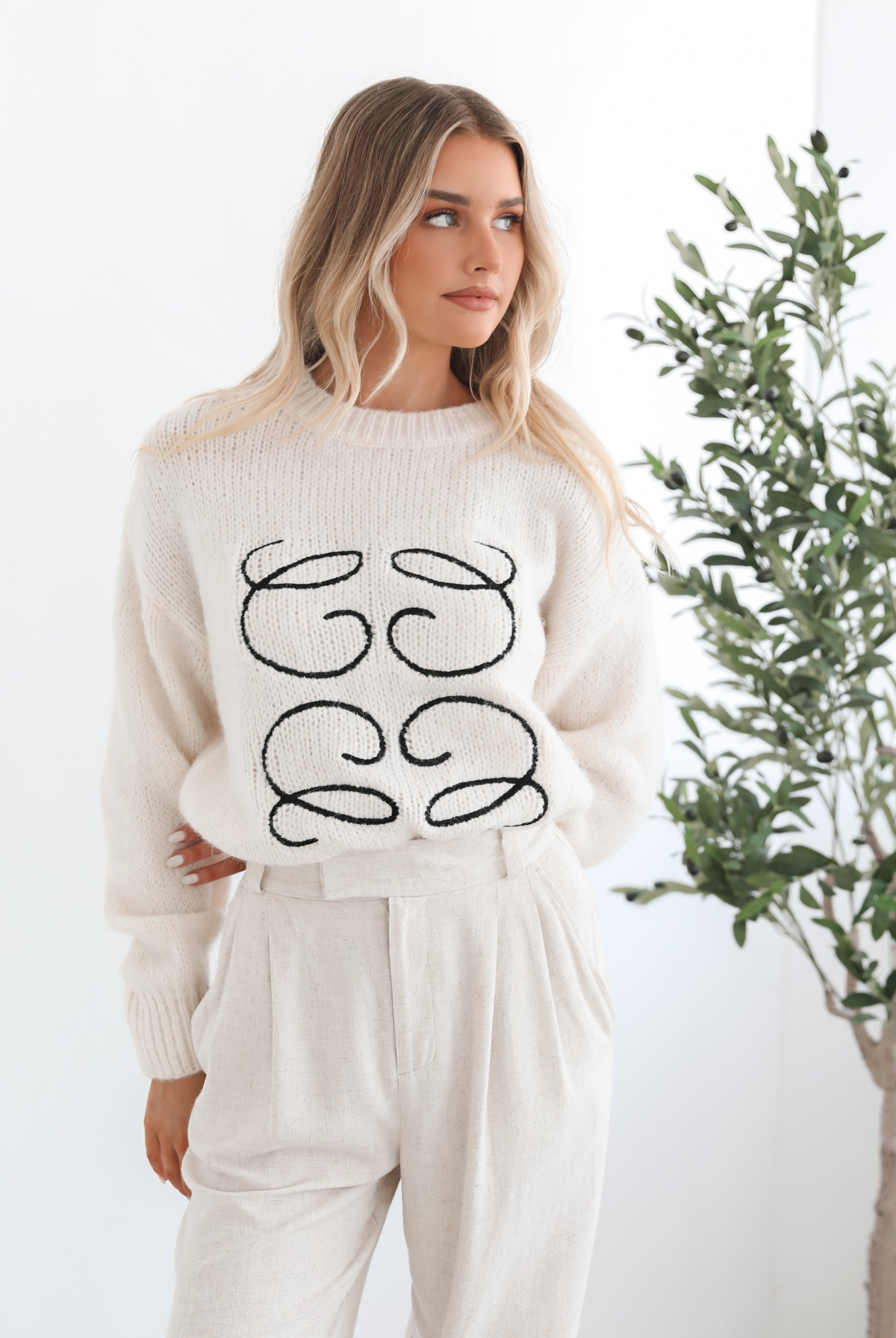 A woman with long, wavy blonde hair stands indoors near a leafy potted plant, wearing a cream-colored sweater with a black abstract design on the front and high-waisted cream trousers. She is gazing to the side with a calm expression, and the minimalist white background enhances the clean, modern look.