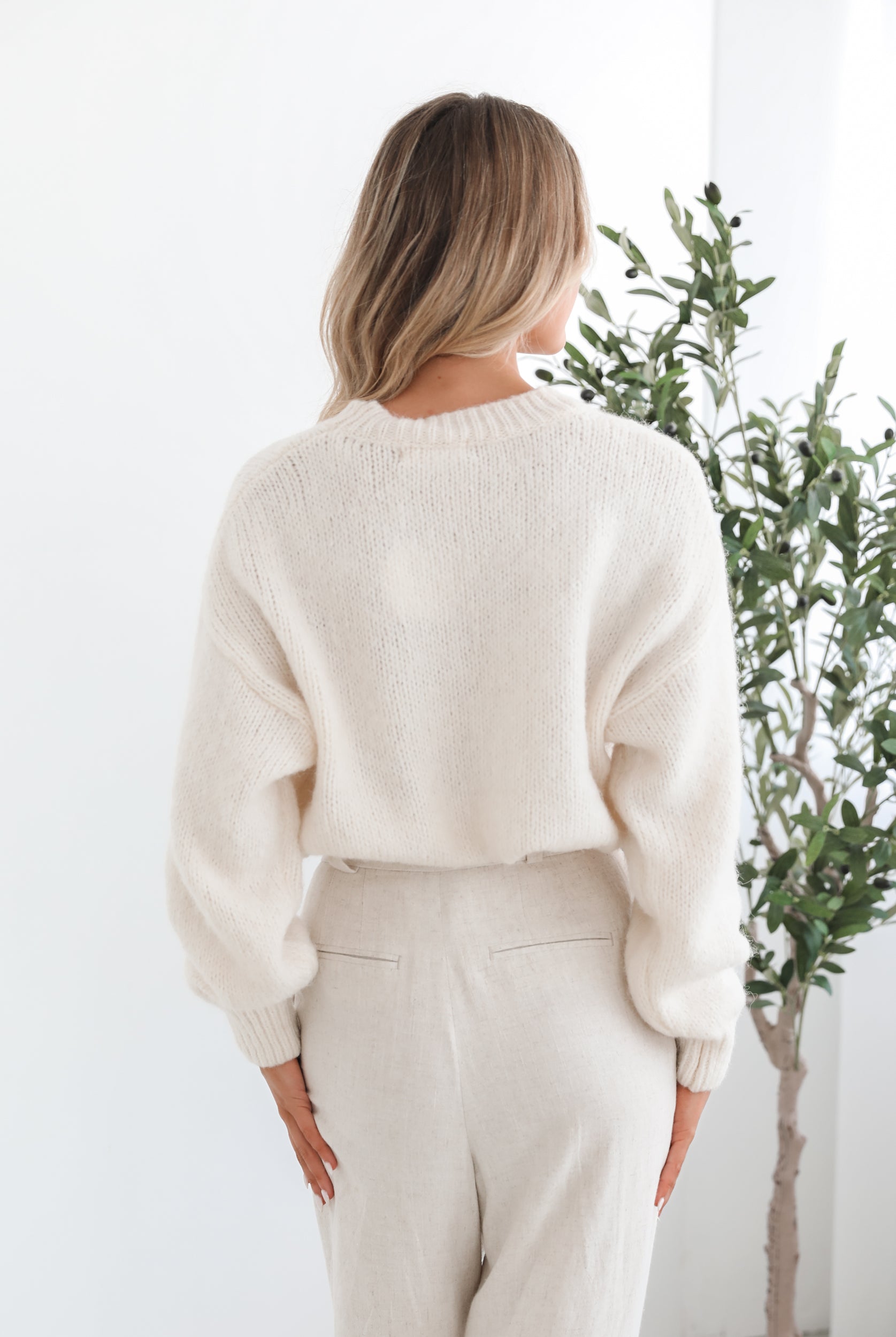 A woman with long, wavy blonde hair stands indoors near a leafy potted plant, wearing a cream-colored sweater with a black abstract design on the front and high-waisted cream trousers. She is gazing to the side with a calm expression, and the minimalist white background enhances the clean, modern look.