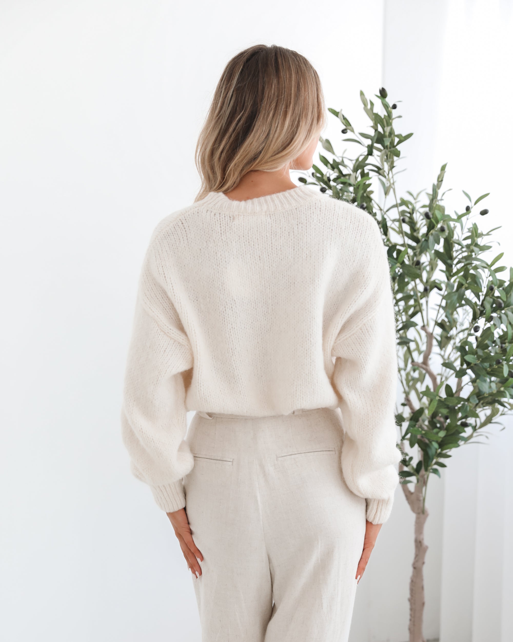 A woman with long, wavy blonde hair stands indoors near a leafy potted plant, wearing a cream-colored sweater with a black abstract design on the front and high-waisted cream trousers. She is gazing to the side with a calm expression, and the minimalist white background enhances the clean, modern look.