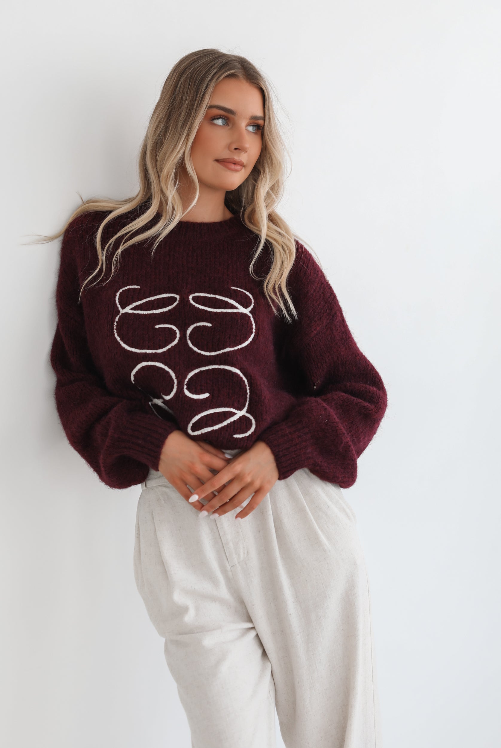A woman with long, wavy blonde hair leans casually against a white wall, wearing a burgundy sweater with a white abstract design on the front and high-waisted cream trousers. She has her hands in her pockets and gazes confidently at the camera, set against a clean, minimalist background.