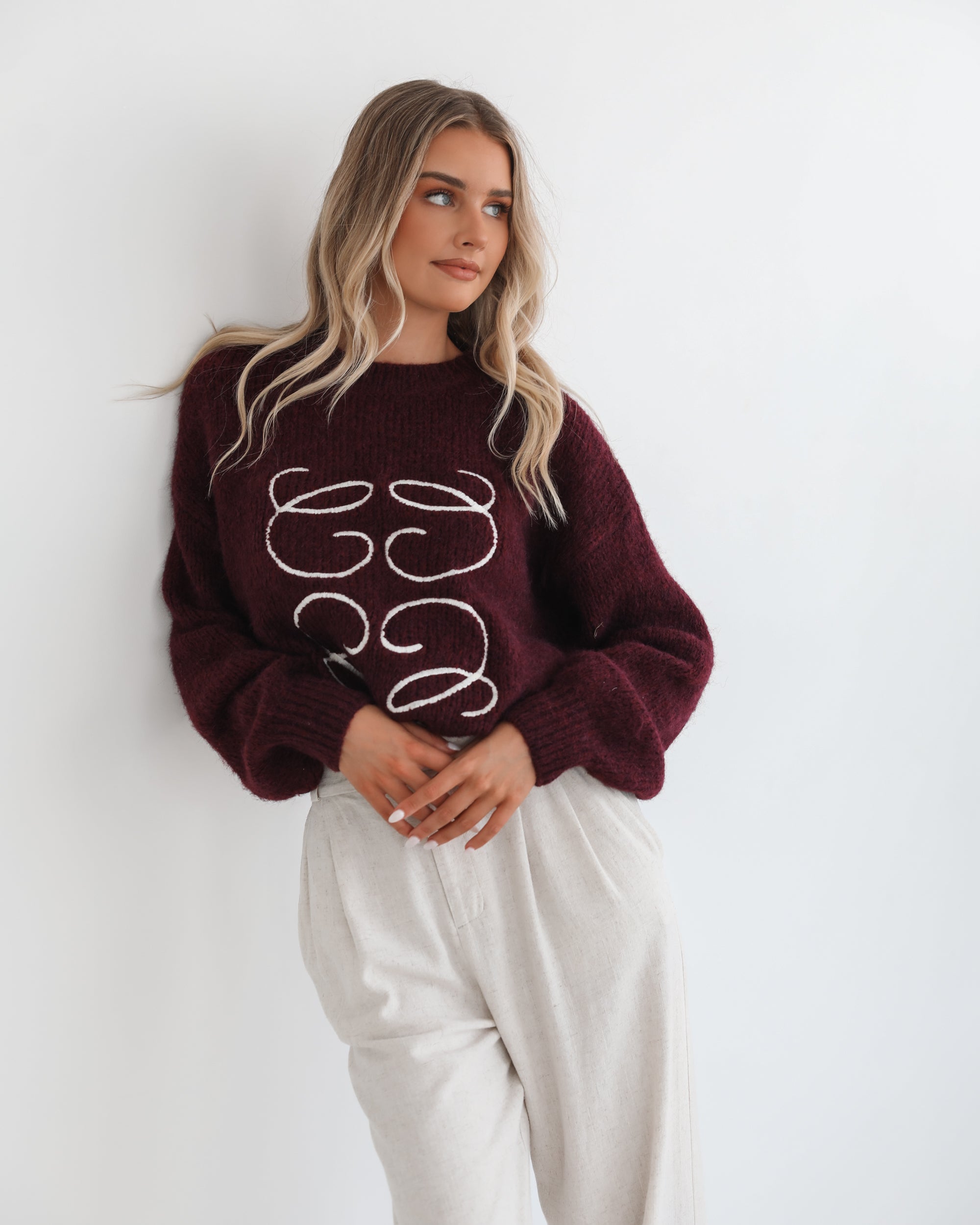 A woman with long, wavy blonde hair leans casually against a white wall, wearing a burgundy sweater with a white abstract design on the front and high-waisted cream trousers. She has her hands in her pockets and gazes confidently at the camera, set against a clean, minimalist background.