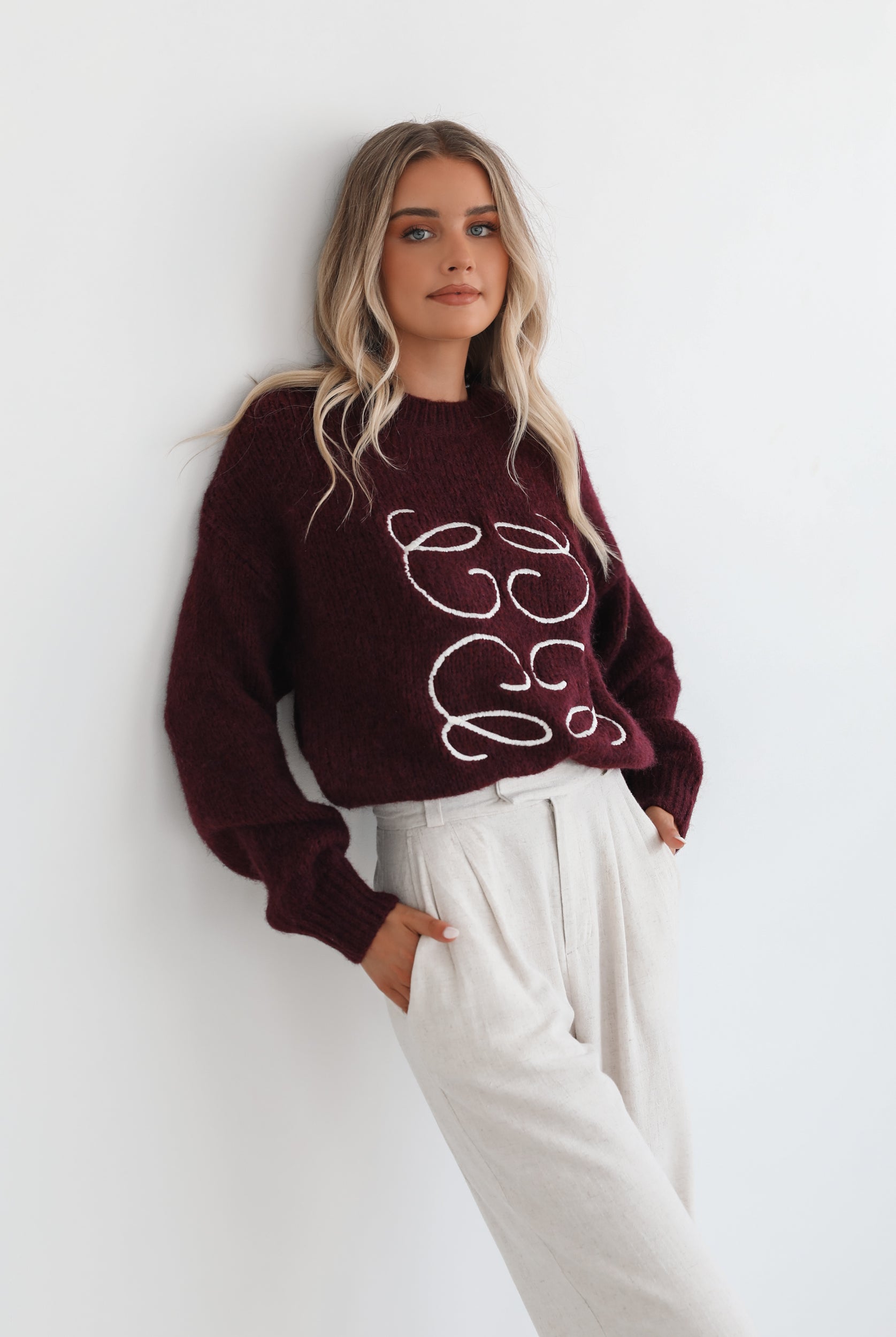 A woman with long, wavy blonde hair leans casually against a white wall, wearing a burgundy sweater with a white abstract design on the front and high-waisted cream trousers. She has her hands in her pockets and gazes confidently at the camera, set against a clean, minimalist background.