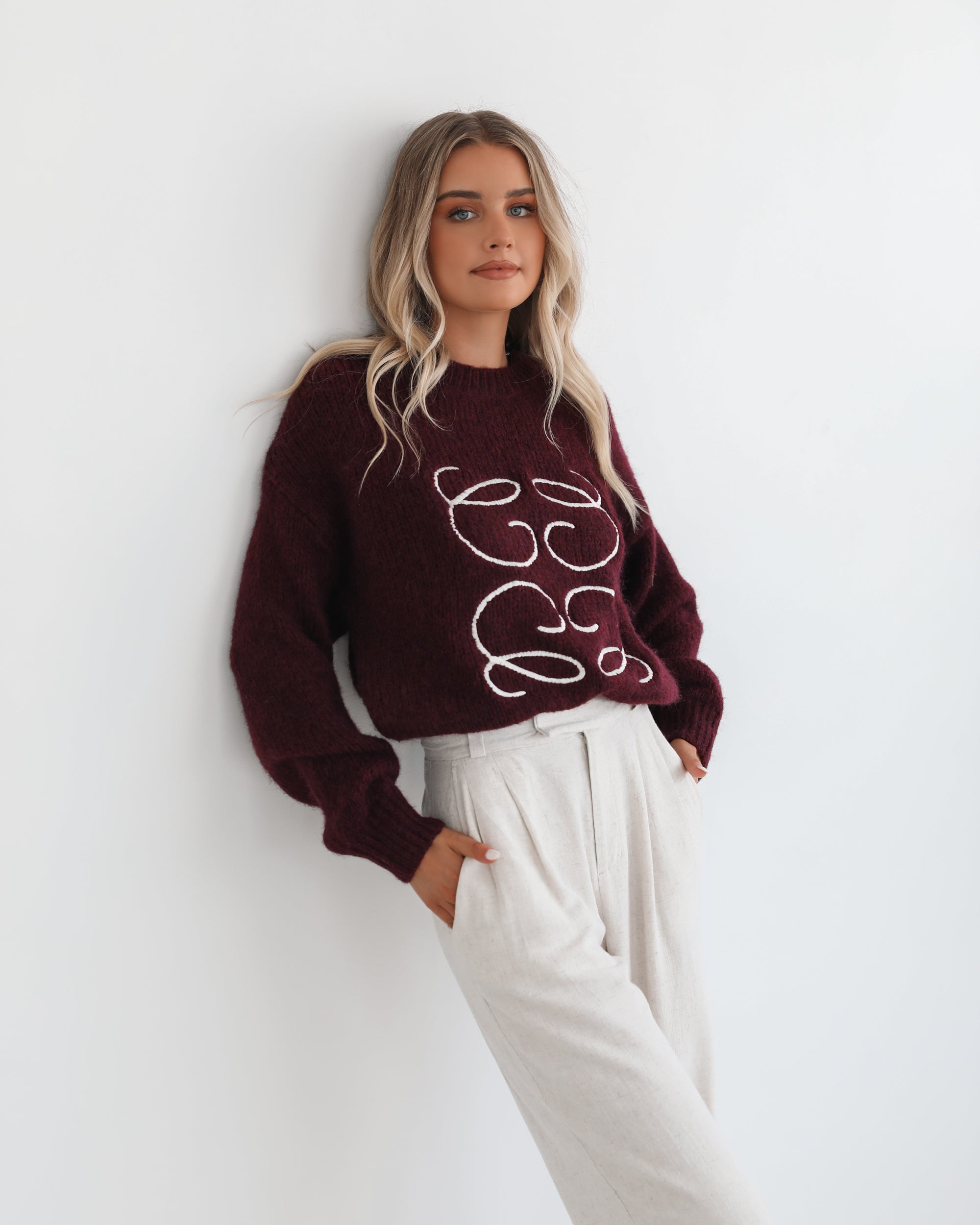 A woman with long, wavy blonde hair leans casually against a white wall, wearing a burgundy sweater with a white abstract design on the front and high-waisted cream trousers. She has her hands in her pockets and gazes confidently at the camera, set against a clean, minimalist background.