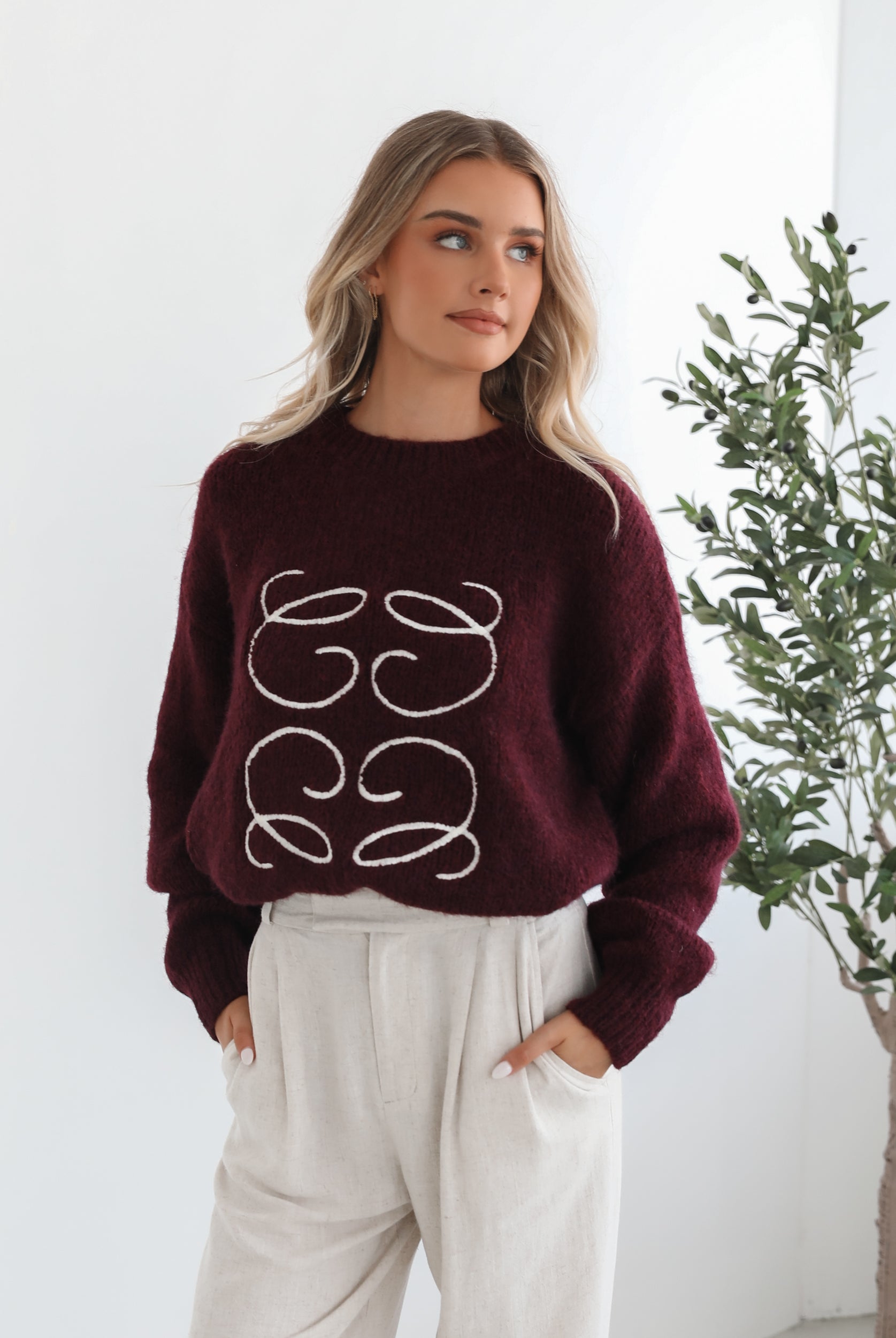 A woman with long, wavy blonde hair leans casually against a white wall, wearing a burgundy sweater with a white abstract design on the front and high-waisted cream trousers. She has her hands in her pockets and gazes confidently at the camera, set against a clean, minimalist background.