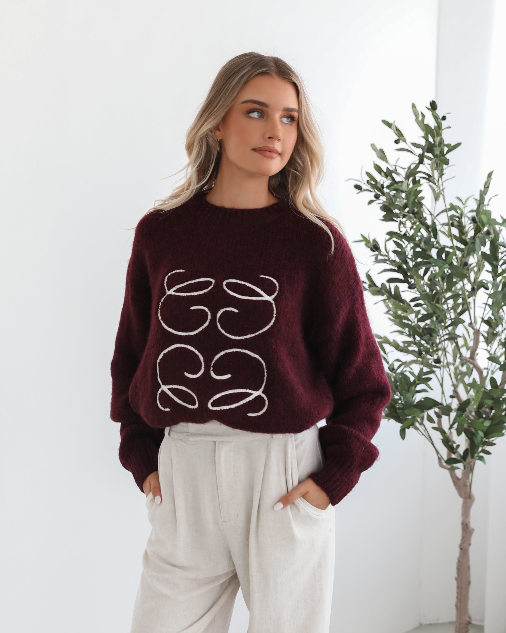 A woman with long, wavy blonde hair leans casually against a white wall, wearing a burgundy sweater with a white abstract design on the front and high-waisted cream trousers. She has her hands in her pockets and gazes confidently at the camera, set against a clean, minimalist background.