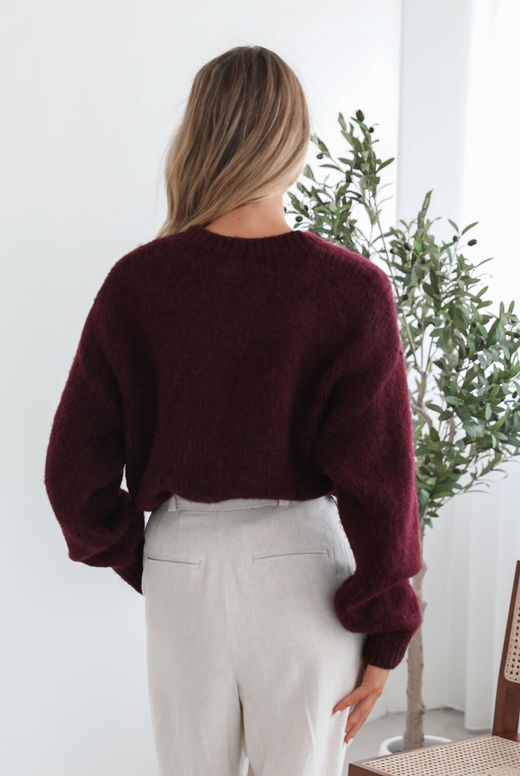 A woman with long, wavy blonde hair leans casually against a white wall, wearing a burgundy sweater with a white abstract design on the front and high-waisted cream trousers. She has her hands in her pockets and gazes confidently at the camera, set against a clean, minimalist background.