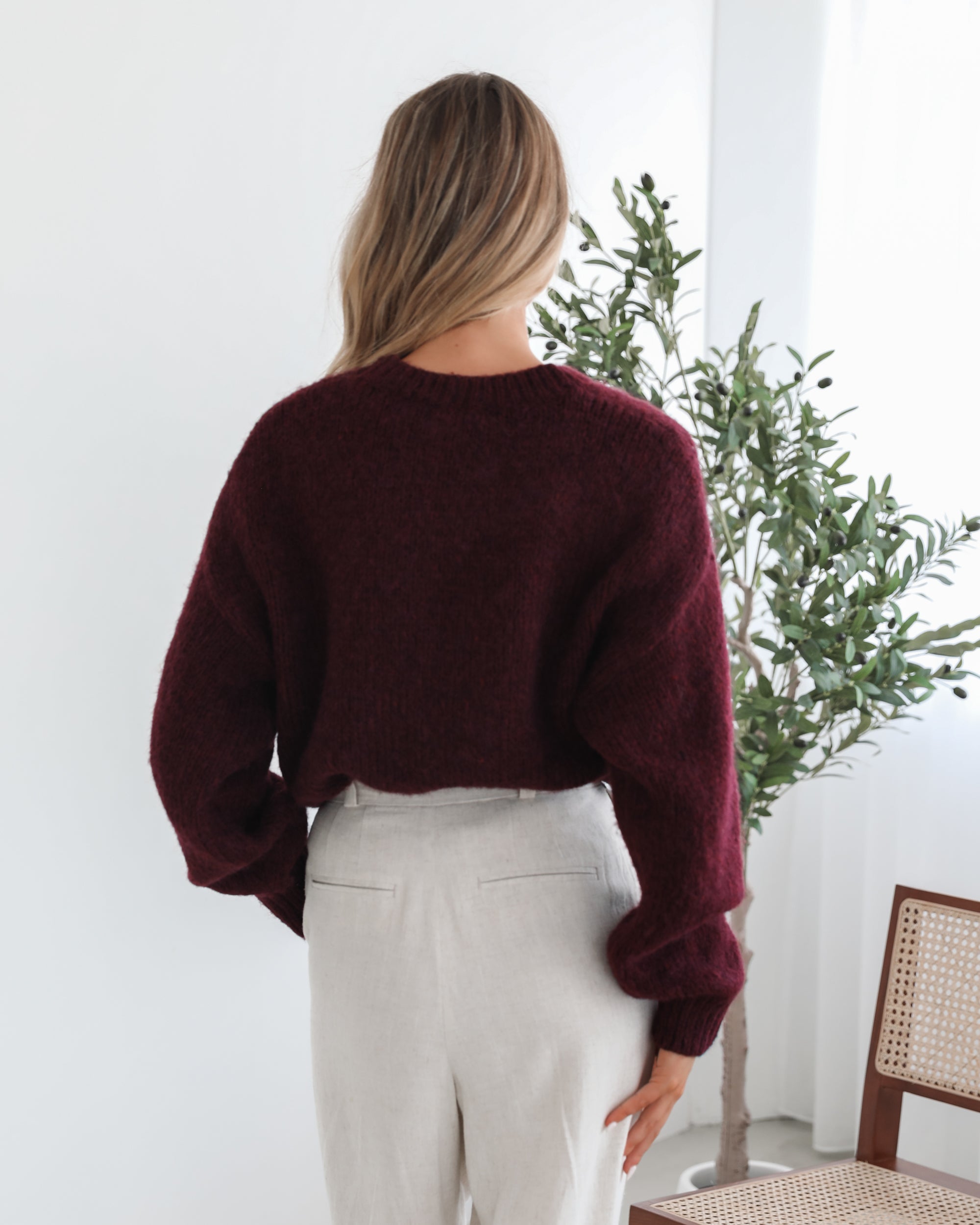A woman with long, wavy blonde hair leans casually against a white wall, wearing a burgundy sweater with a white abstract design on the front and high-waisted cream trousers. She has her hands in her pockets and gazes confidently at the camera, set against a clean, minimalist background.