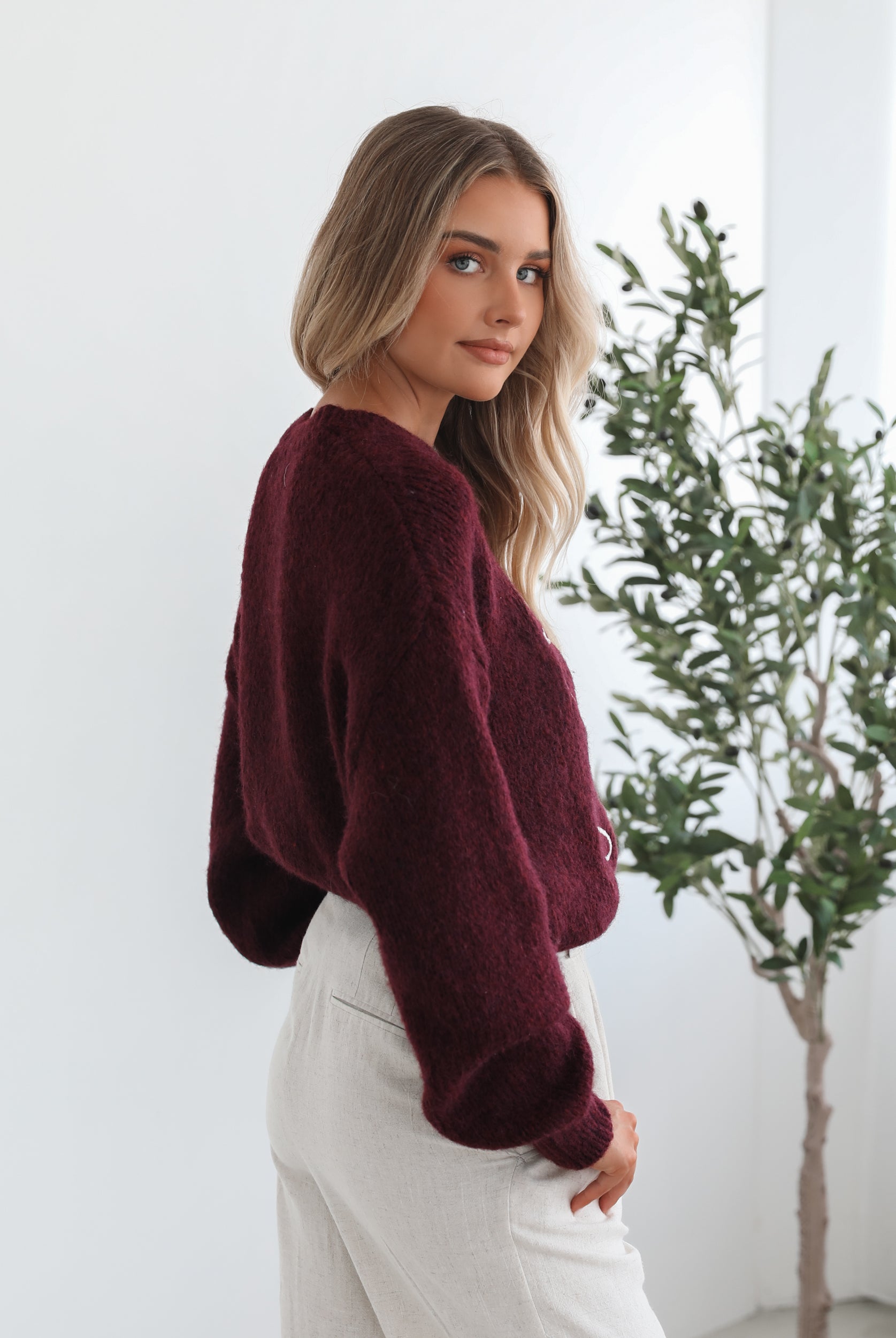 A woman with long, wavy blonde hair leans casually against a white wall, wearing a burgundy sweater with a white abstract design on the front and high-waisted cream trousers. She has her hands in her pockets and gazes confidently at the camera, set against a clean, minimalist background.