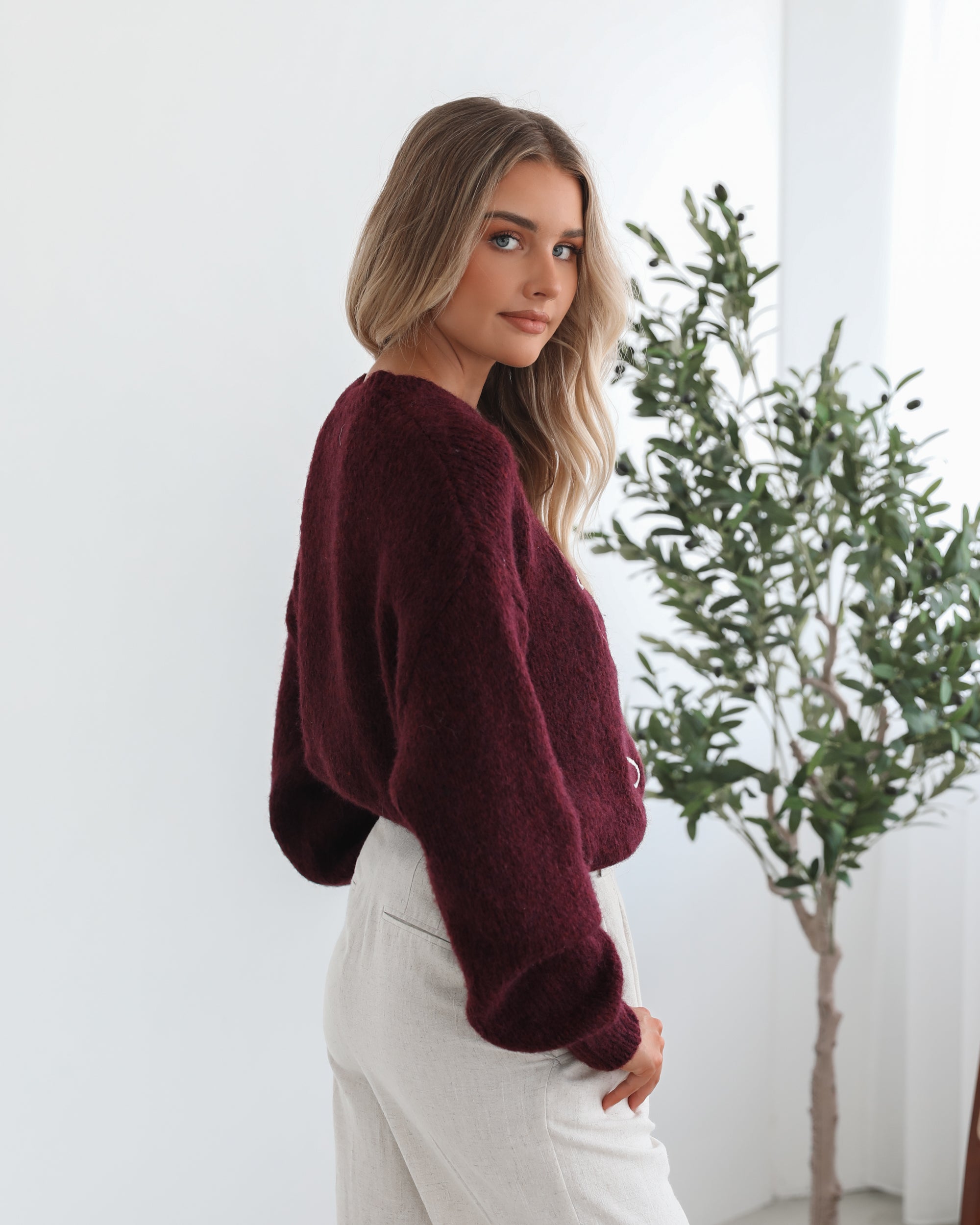 A woman with long, wavy blonde hair leans casually against a white wall, wearing a burgundy sweater with a white abstract design on the front and high-waisted cream trousers. She has her hands in her pockets and gazes confidently at the camera, set against a clean, minimalist background.