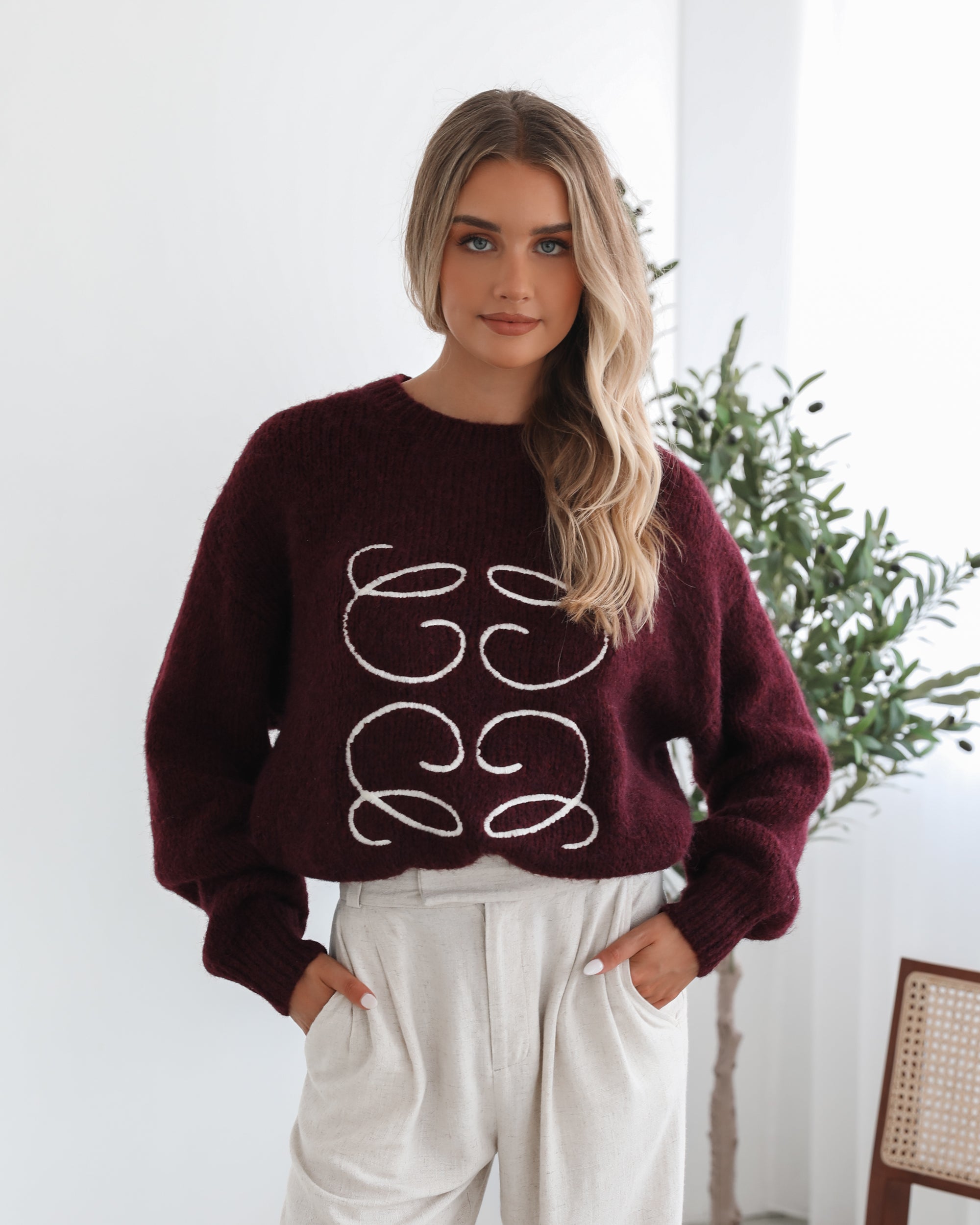 A woman with long, wavy blonde hair leans casually against a white wall, wearing a burgundy sweater with a white abstract design on the front and high-waisted cream trousers. She has her hands in her pockets and gazes confidently at the camera, set against a clean, minimalist background.