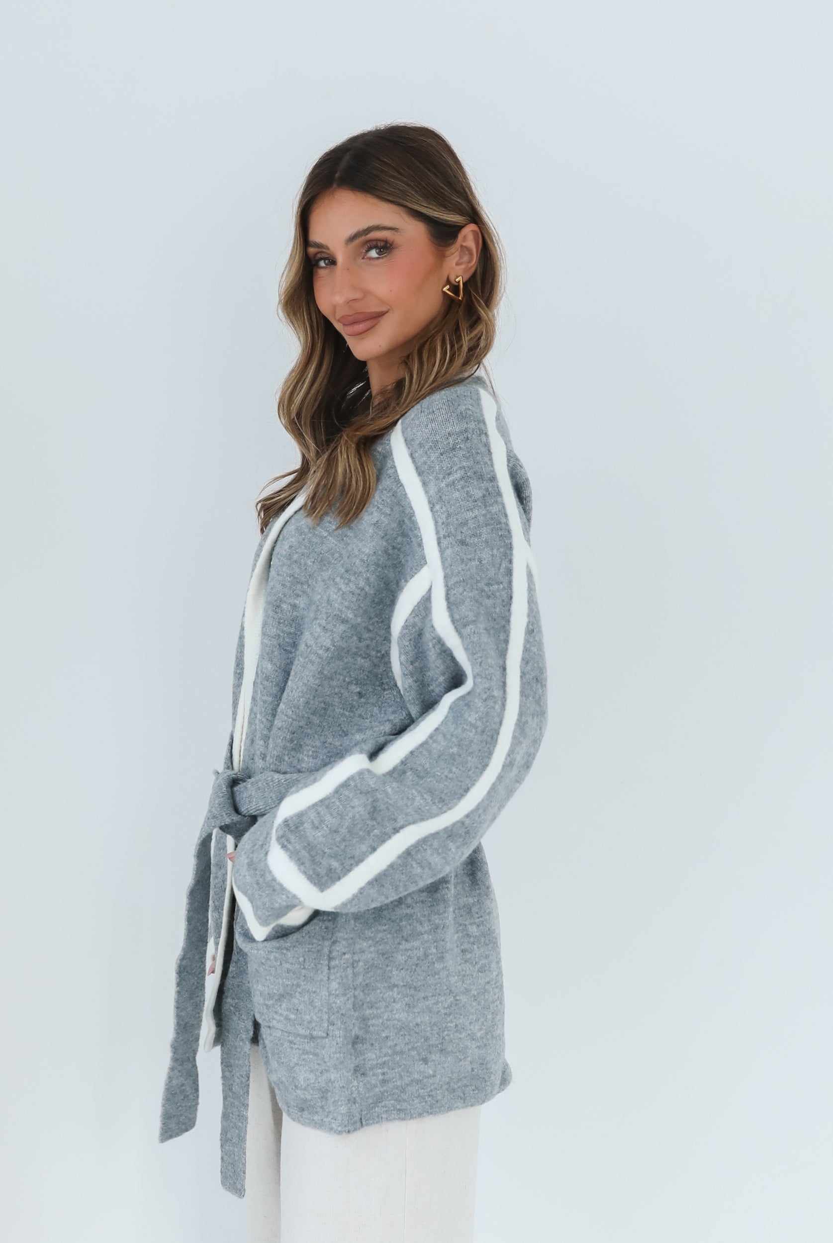 A smiling woman with long, wavy brown hair stands against a plain white background, wearing a cozy grey cardigan with white trim over a white top and loose-fitting cream pants. She has her hands gently clasped in front of her, giving a relaxed and cheerful vibe. A small sprig of greenery is visible to the left.