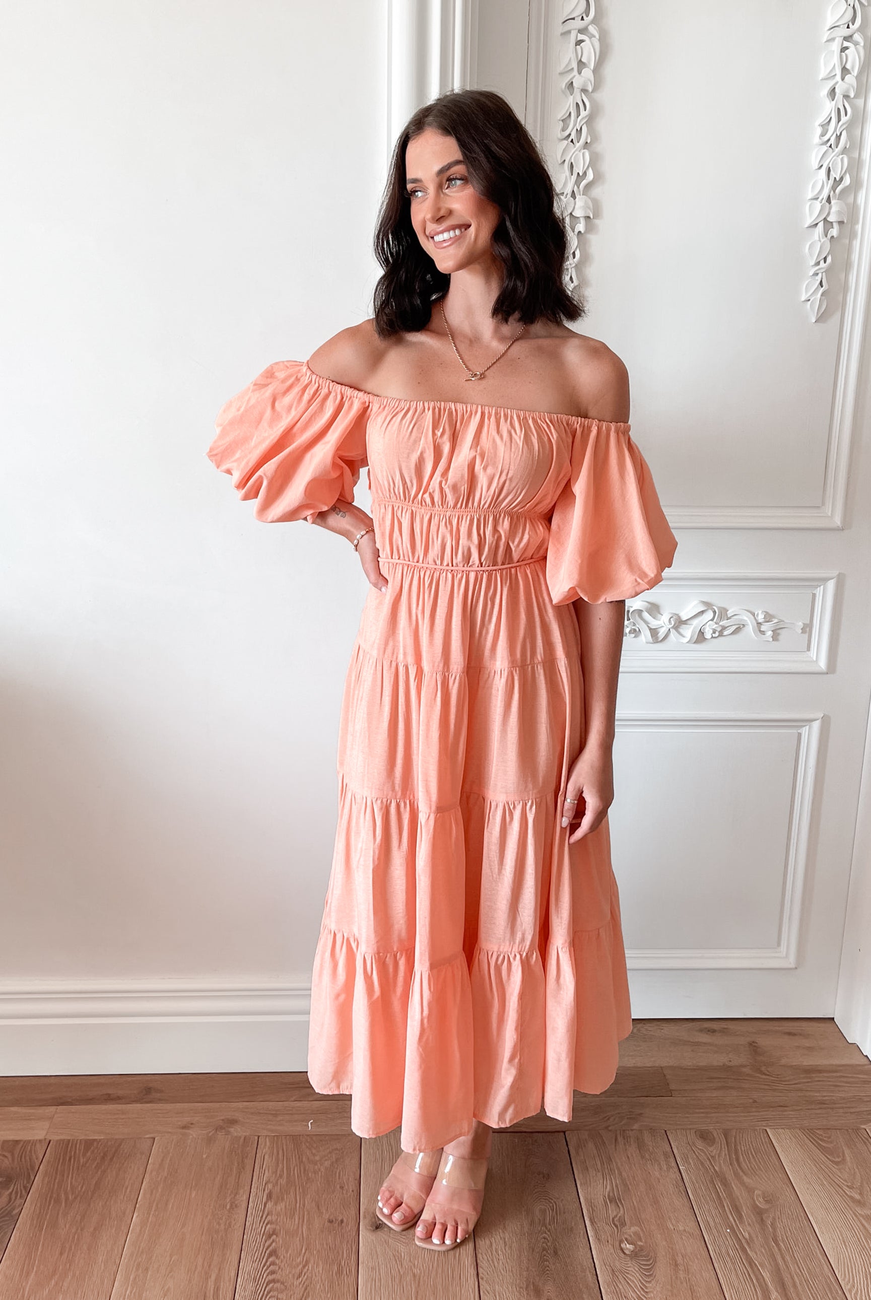 A woman with shoulder-length dark hair stands in a beautifully decorated room with white ornate wall paneling and wooden flooring, wearing an off-the-shoulder peach midi dress with puffed sleeves and a tiered skirt. She is smiling and gently holding one side of her dress, giving a soft and elegant pose.