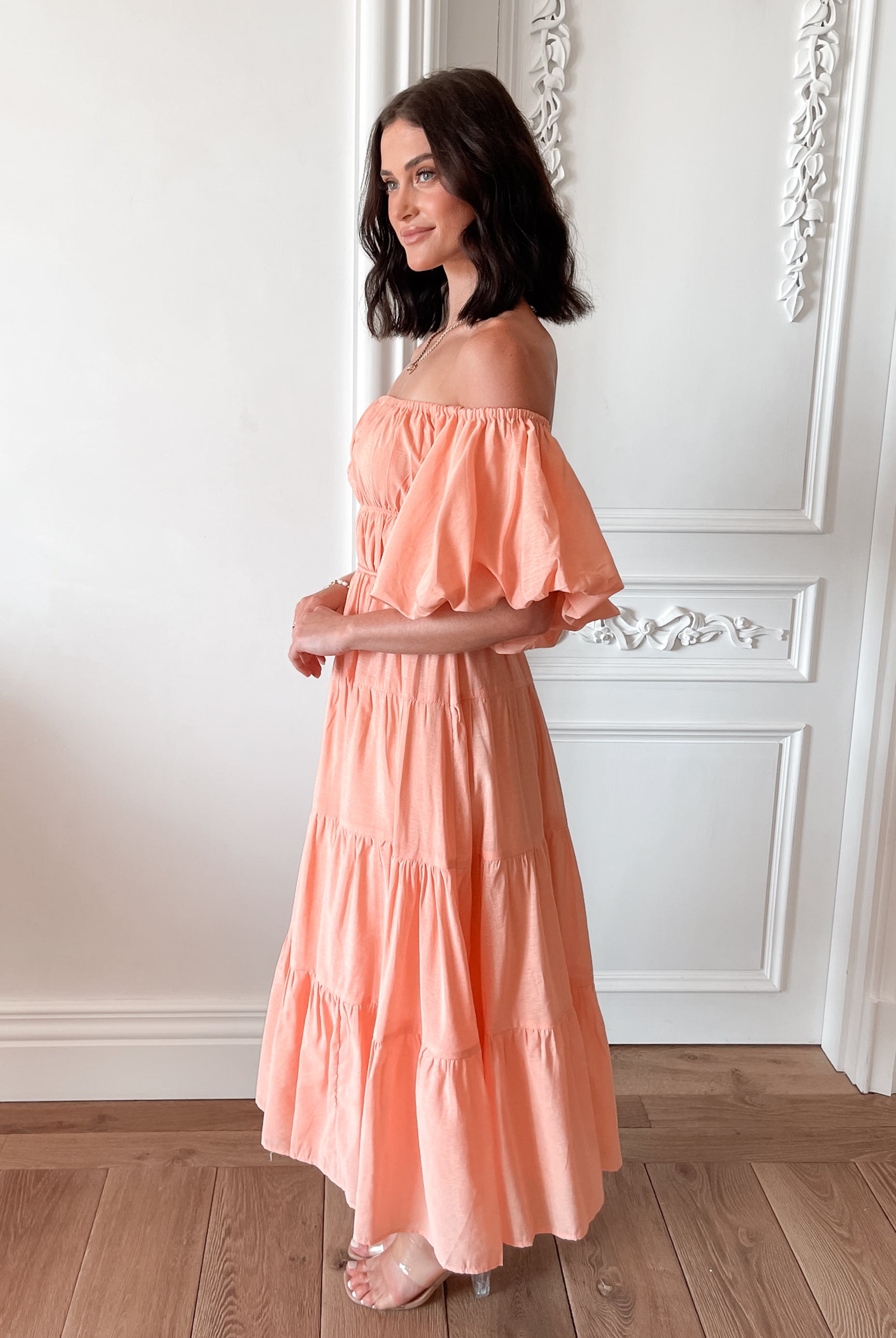 A woman with shoulder-length dark hair stands in a beautifully decorated room with white ornate wall paneling and wooden flooring, wearing an off-the-shoulder peach midi dress with puffed sleeves and a tiered skirt. She is smiling and gently holding one side of her dress, giving a soft and elegant pose.