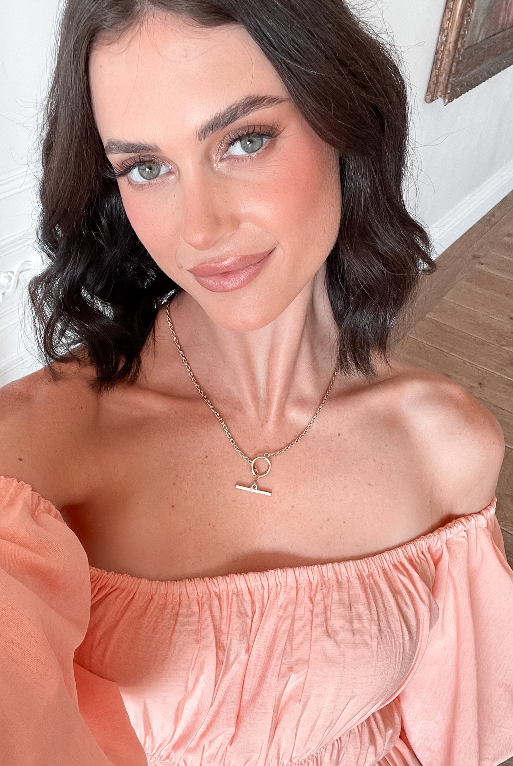 A woman with shoulder-length dark hair stands in a beautifully decorated room with white ornate wall paneling and wooden flooring, wearing an off-the-shoulder peach midi dress with puffed sleeves and a tiered skirt. She is smiling and gently holding one side of her dress, giving a soft and elegant pose.