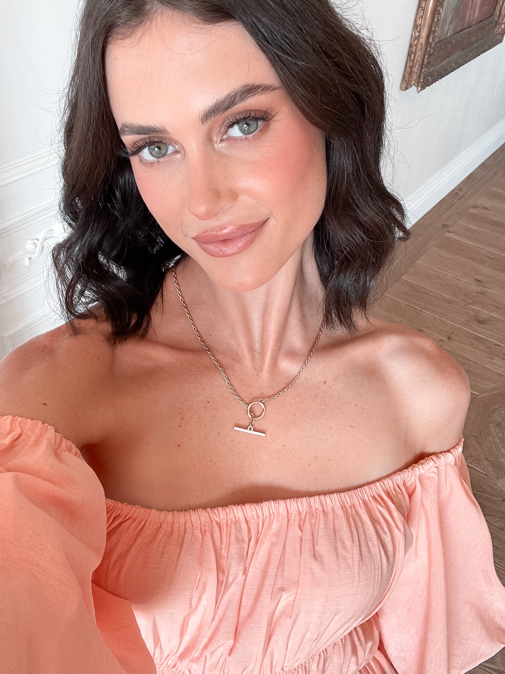 A woman with shoulder-length dark hair stands in a beautifully decorated room with white ornate wall paneling and wooden flooring, wearing an off-the-shoulder peach midi dress with puffed sleeves and a tiered skirt. She is smiling and gently holding one side of her dress, giving a soft and elegant pose.