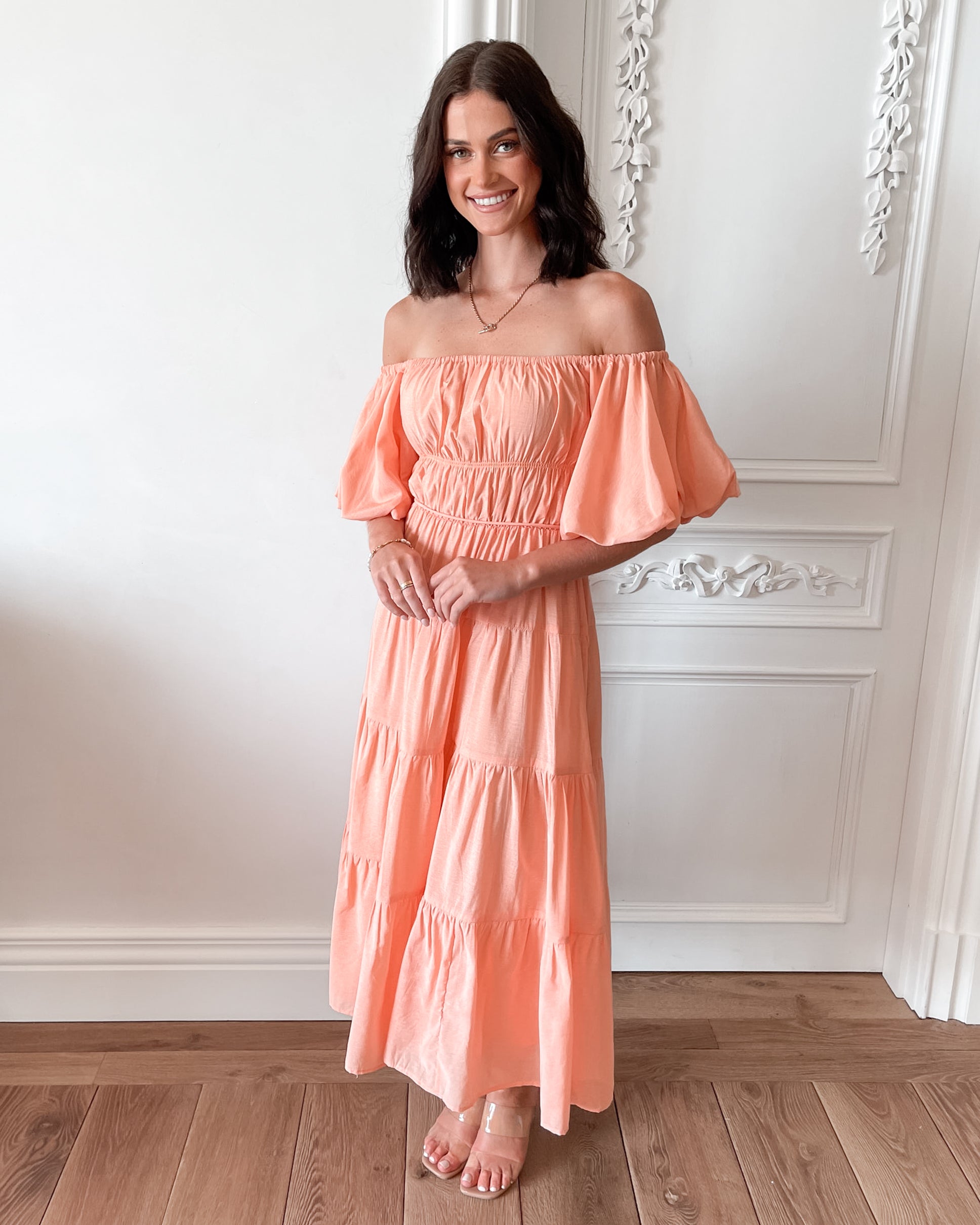 A woman with shoulder-length dark hair stands in a beautifully decorated room with white ornate wall paneling and wooden flooring, wearing an off-the-shoulder peach midi dress with puffed sleeves and a tiered skirt. She is smiling and gently holding one side of her dress, giving a soft and elegant pose.