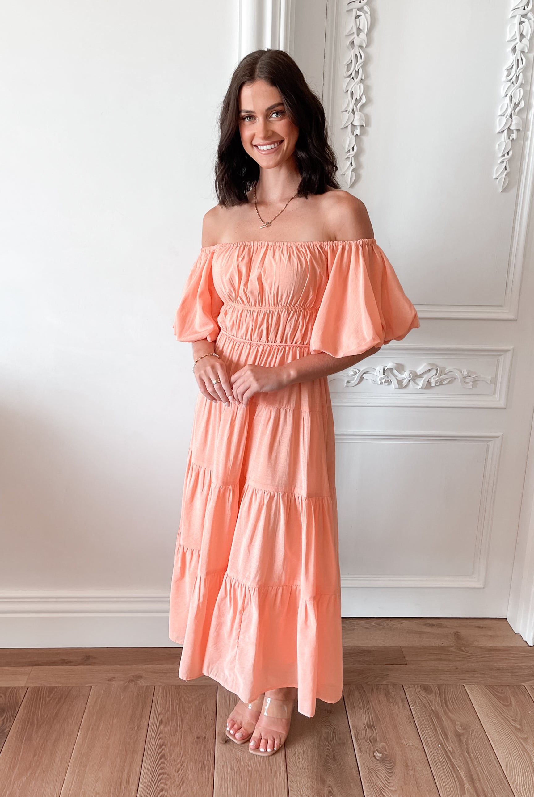 A woman with shoulder-length dark hair stands in a beautifully decorated room with white ornate wall paneling and wooden flooring, wearing an off-the-shoulder peach midi dress with puffed sleeves and a tiered skirt. She is smiling and gently holding one side of her dress, giving a soft and elegant pose.