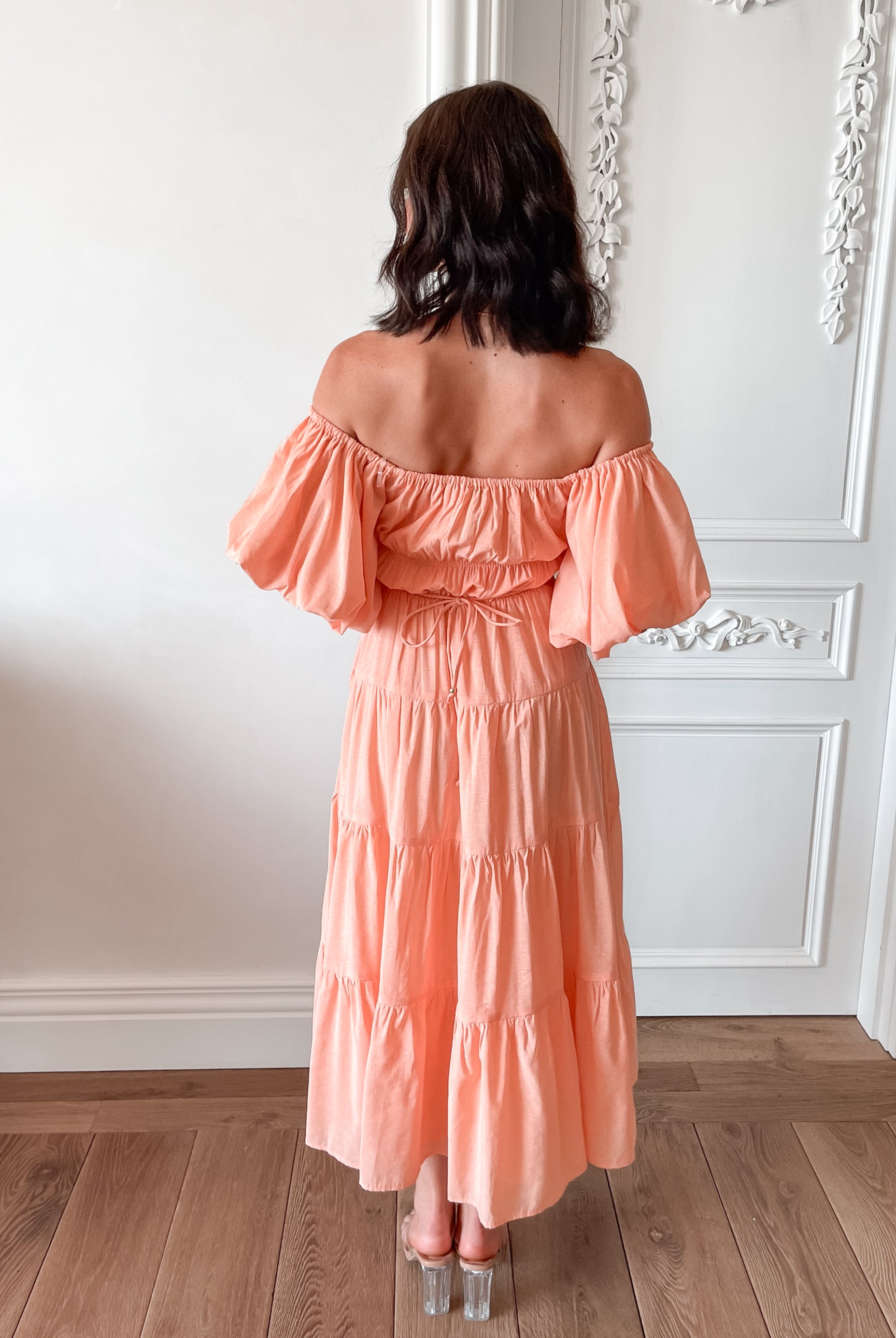 A woman with shoulder-length dark hair stands in a beautifully decorated room with white ornate wall paneling and wooden flooring, wearing an off-the-shoulder peach midi dress with puffed sleeves and a tiered skirt. She is smiling and gently holding one side of her dress, giving a soft and elegant pose.