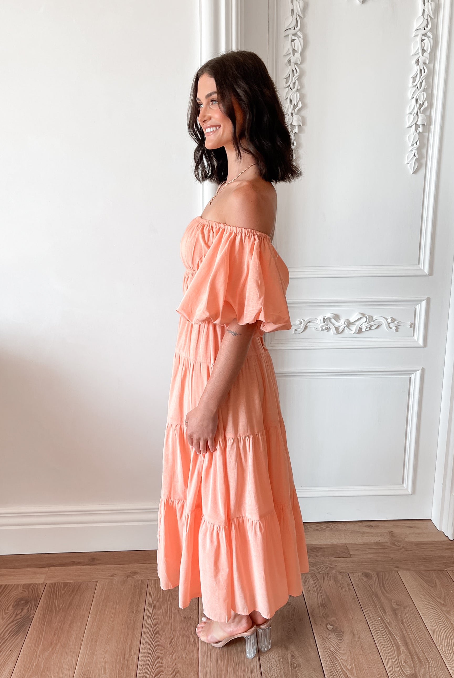 A woman with shoulder-length dark hair stands in a beautifully decorated room with white ornate wall paneling and wooden flooring, wearing an off-the-shoulder peach midi dress with puffed sleeves and a tiered skirt. She is smiling and gently holding one side of her dress, giving a soft and elegant pose.