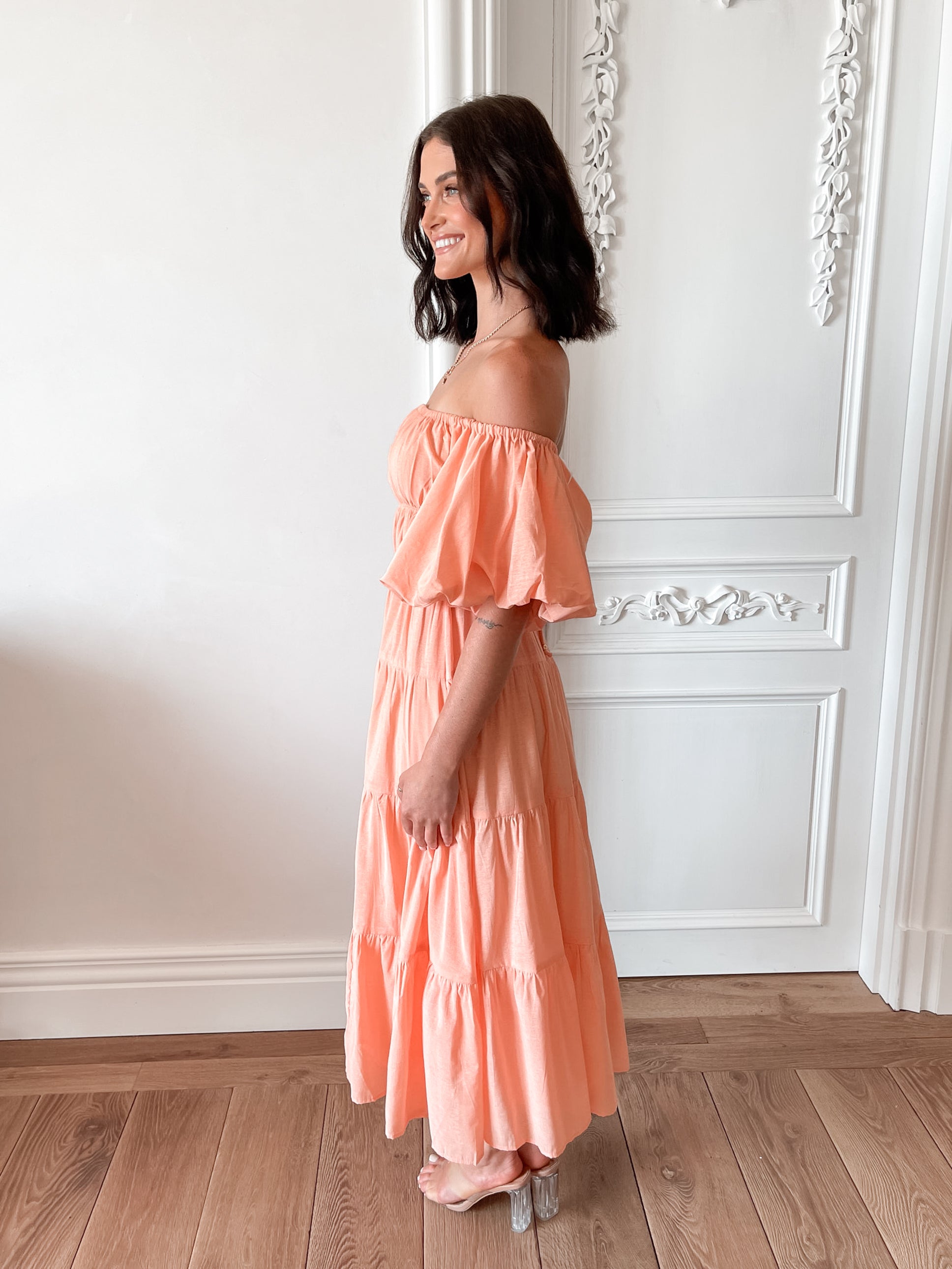 A woman with shoulder-length dark hair stands in a beautifully decorated room with white ornate wall paneling and wooden flooring, wearing an off-the-shoulder peach midi dress with puffed sleeves and a tiered skirt. She is smiling and gently holding one side of her dress, giving a soft and elegant pose.