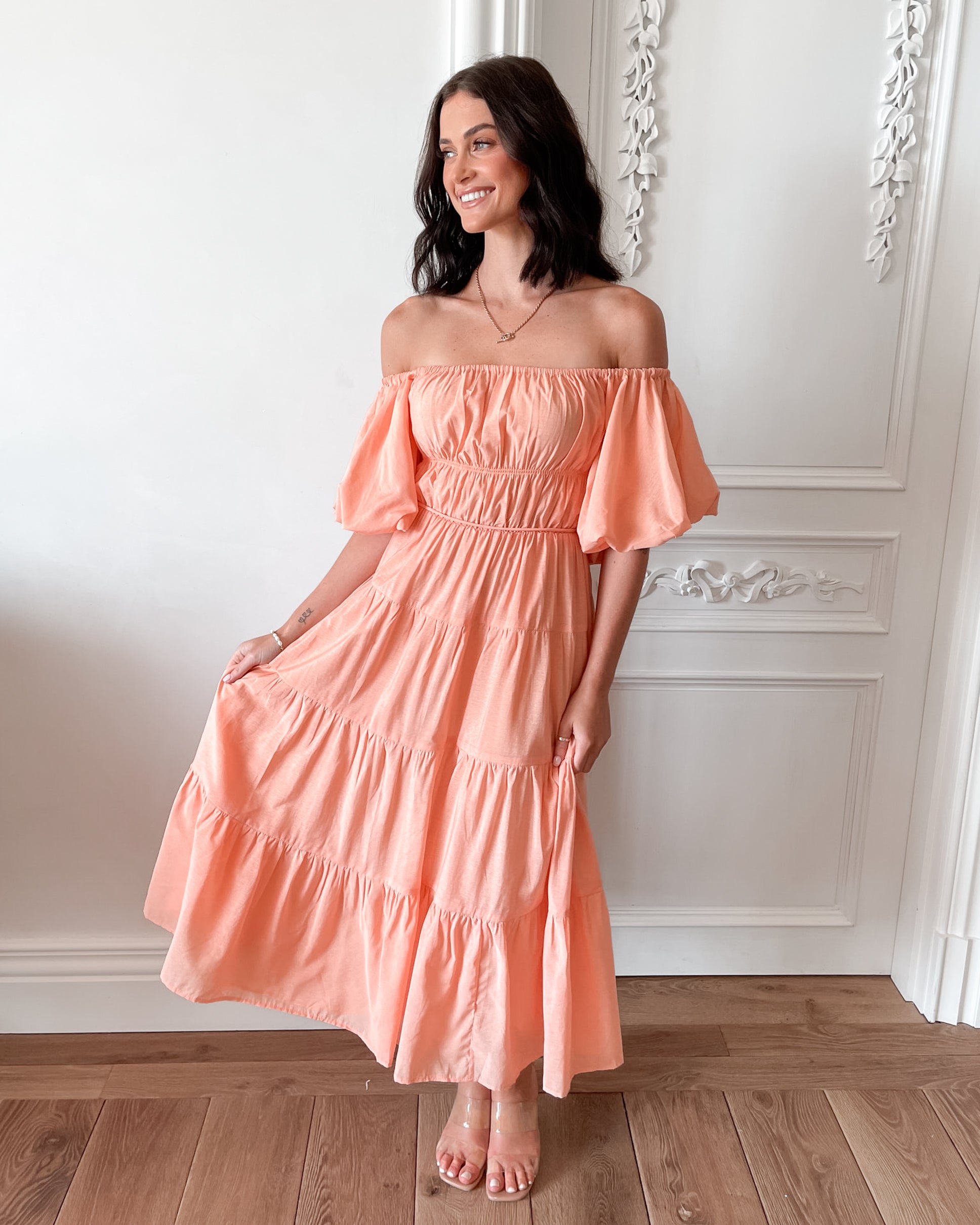 A woman with shoulder-length dark hair stands in a beautifully decorated room with white ornate wall paneling and wooden flooring, wearing an off-the-shoulder peach midi dress with puffed sleeves and a tiered skirt. She is smiling and gently holding one side of her dress, giving a soft and elegant pose.