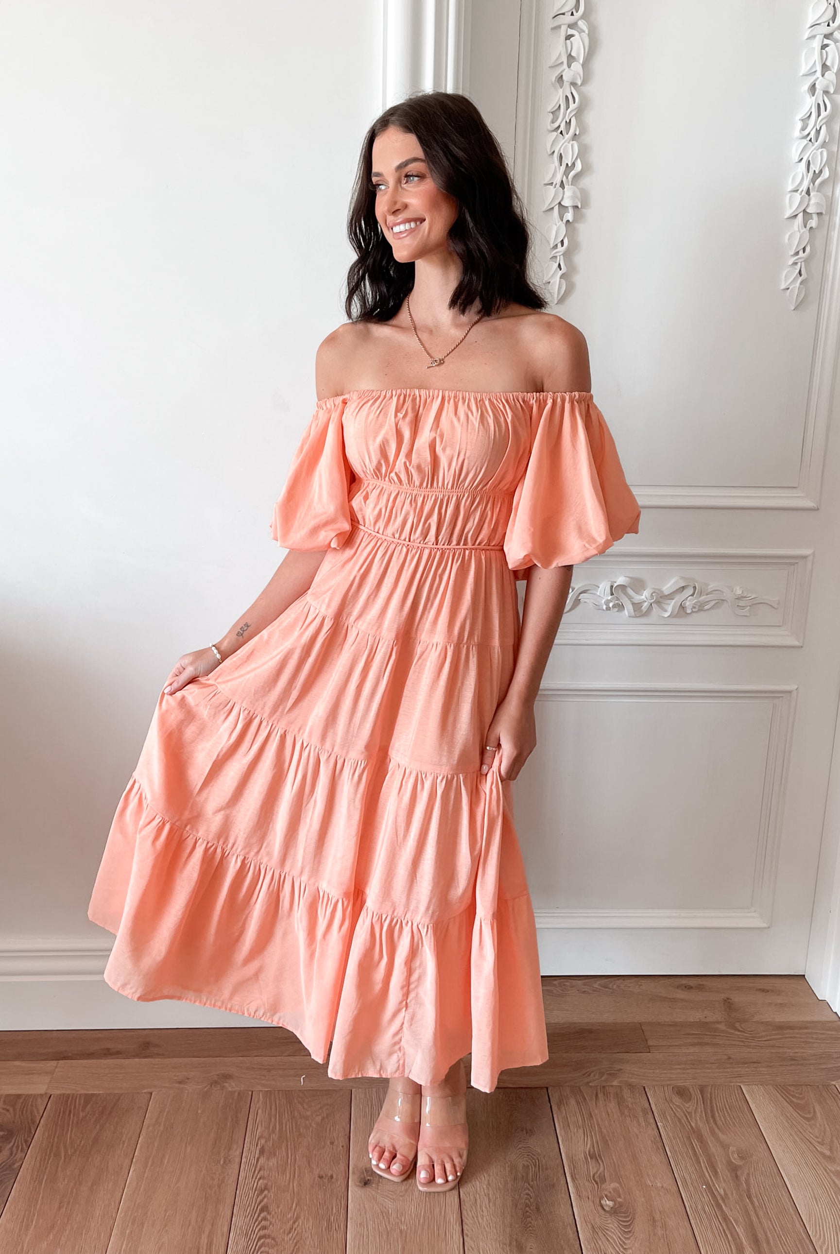 A woman with shoulder-length dark hair stands in a beautifully decorated room with white ornate wall paneling and wooden flooring, wearing an off-the-shoulder peach midi dress with puffed sleeves and a tiered skirt. She is smiling and gently holding one side of her dress, giving a soft and elegant pose.