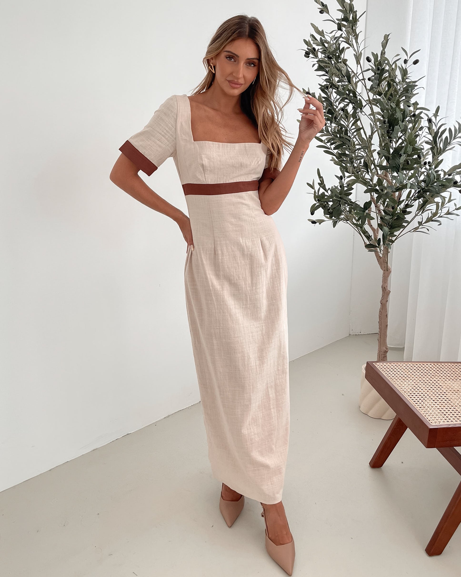 A woman with long, wavy blonde hair poses indoors in a minimalist room with white walls and a potted plant. She is wearing a light beige short-sleeve dress with brown trim details at the sleeves and waist, featuring a square neckline and a slim fit. She completes the look with pointed nude heels and a soft, confident expression.
