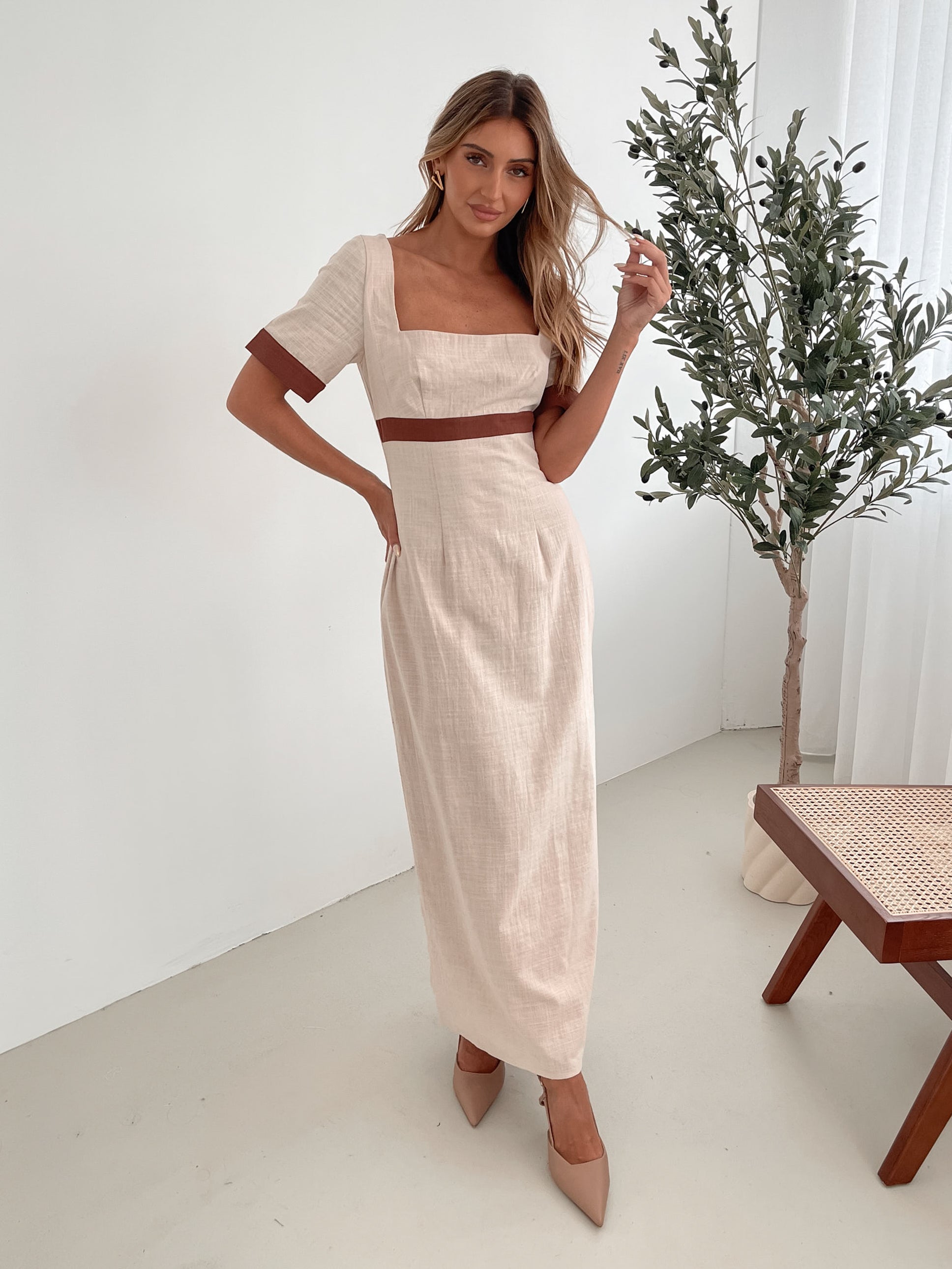 A woman with long, wavy blonde hair poses indoors in a minimalist room with white walls and a potted plant. She is wearing a light beige short-sleeve dress with brown trim details at the sleeves and waist, featuring a square neckline and a slim fit. She completes the look with pointed nude heels and a soft, confident expression.