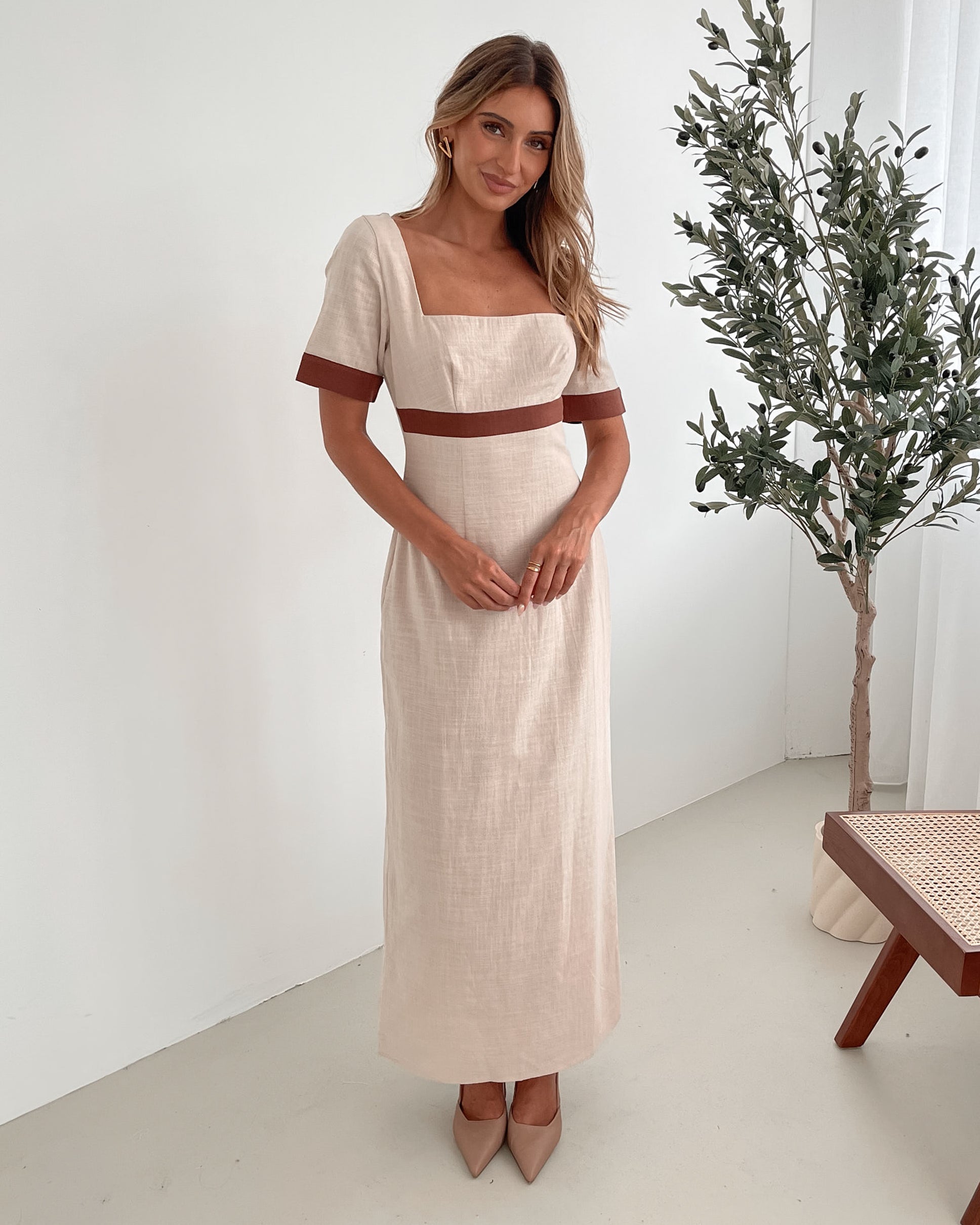 A woman with long, wavy blonde hair poses indoors in a minimalist room with white walls and a potted plant. She is wearing a light beige short-sleeve dress with brown trim details at the sleeves and waist, featuring a square neckline and a slim fit. She completes the look with pointed nude heels and a soft, confident expression.