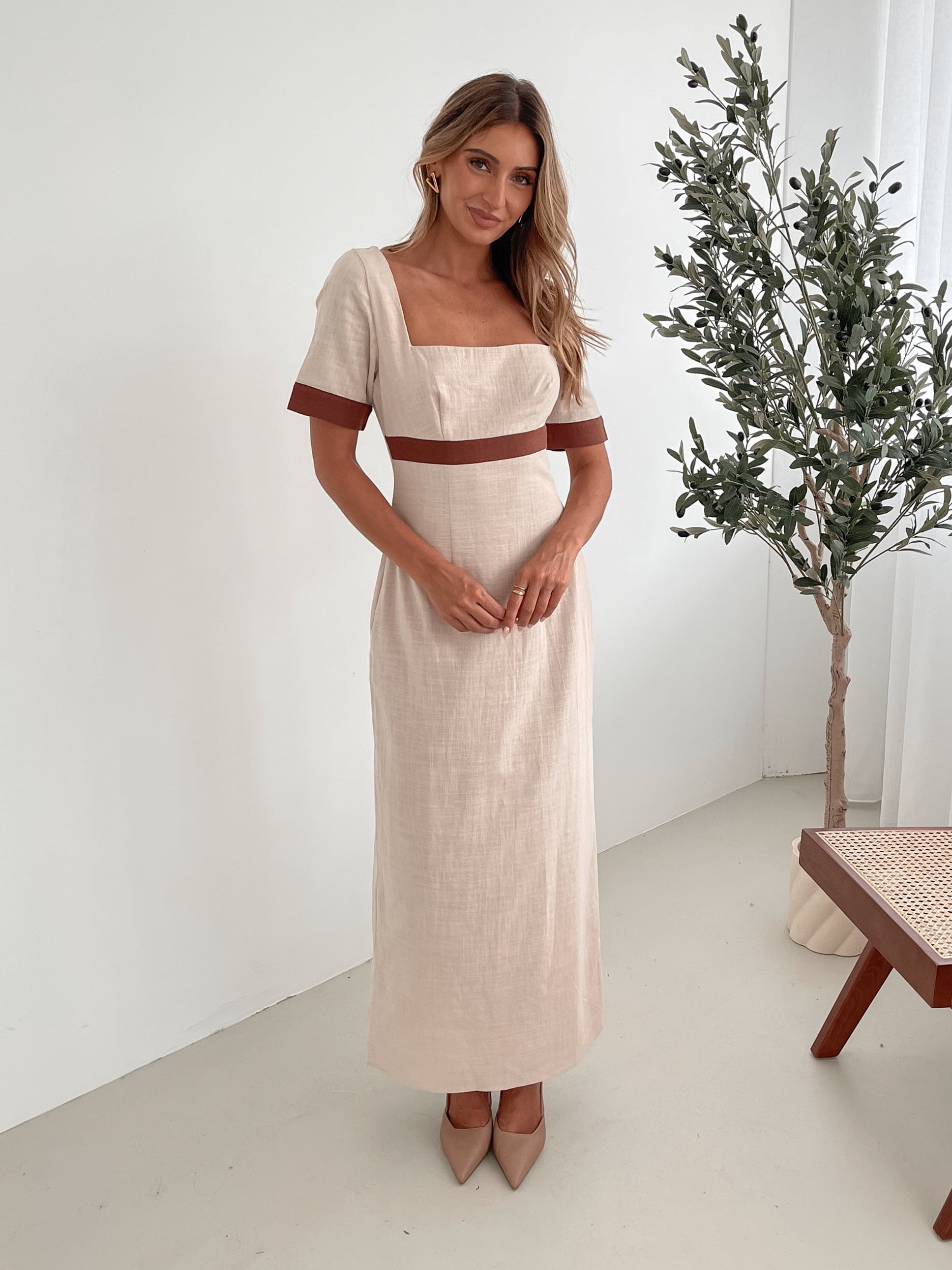 A woman with long, wavy blonde hair poses indoors in a minimalist room with white walls and a potted plant. She is wearing a light beige short-sleeve dress with brown trim details at the sleeves and waist, featuring a square neckline and a slim fit. She completes the look with pointed nude heels and a soft, confident expression.