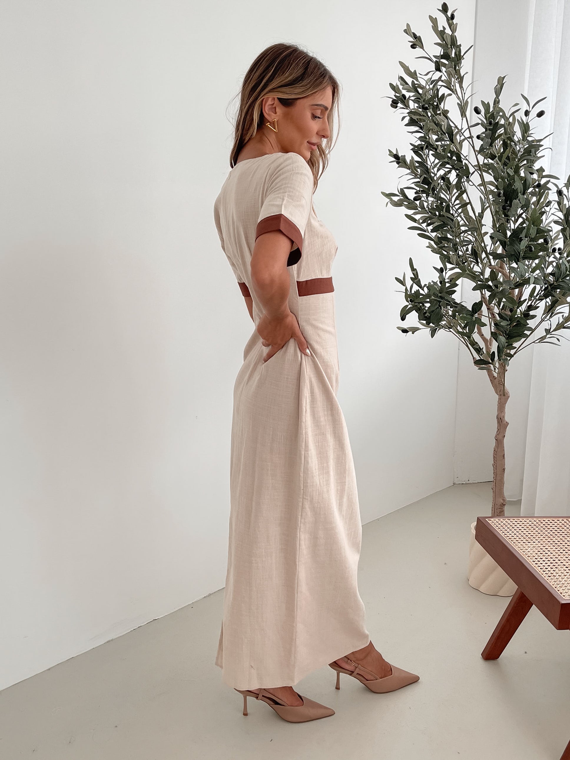 A woman with long, wavy blonde hair poses indoors in a minimalist room with white walls and a potted plant. She is wearing a light beige short-sleeve dress with brown trim details at the sleeves and waist, featuring a square neckline and a slim fit. She completes the look with pointed nude heels and a soft, confident expression.