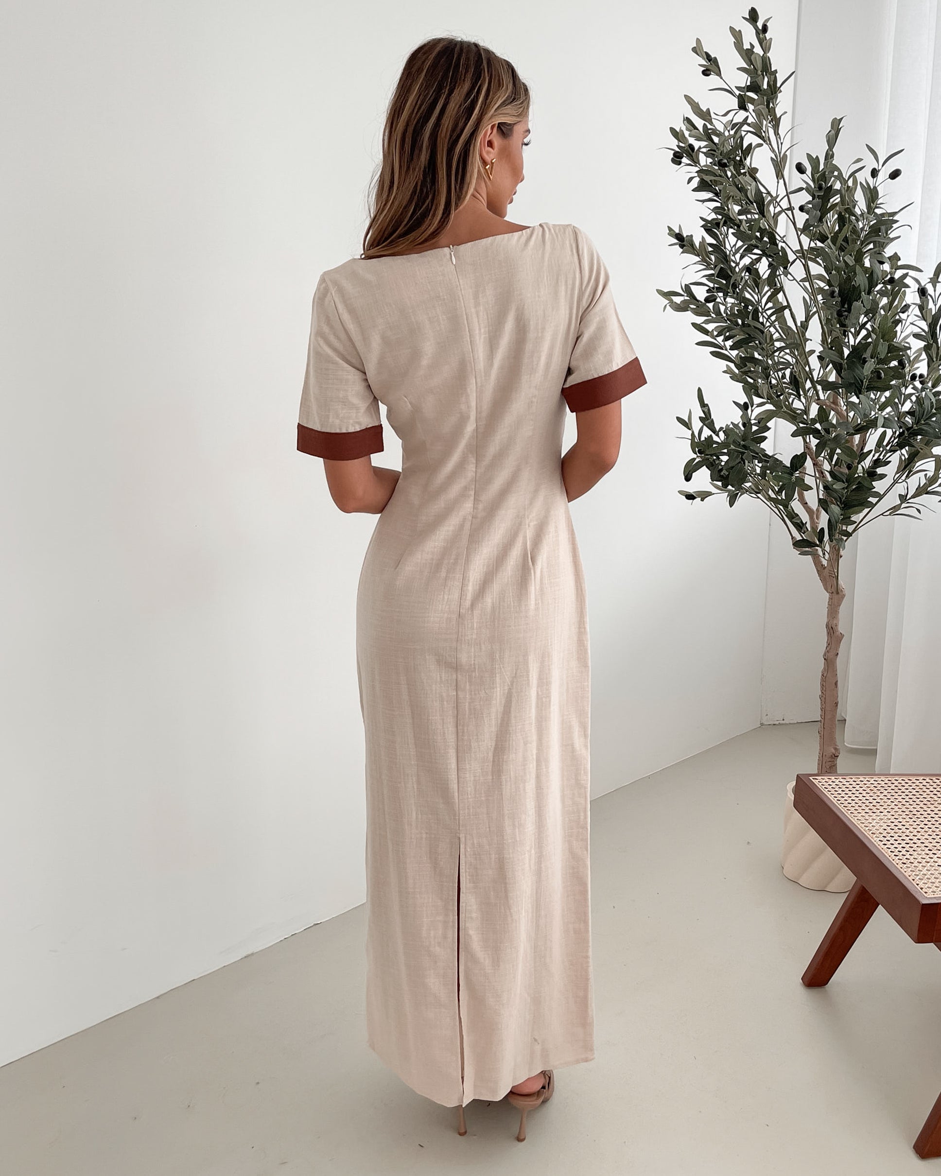 A woman with long, wavy blonde hair poses indoors in a minimalist room with white walls and a potted plant. She is wearing a light beige short-sleeve dress with brown trim details at the sleeves and waist, featuring a square neckline and a slim fit. She completes the look with pointed nude heels and a soft, confident expression.