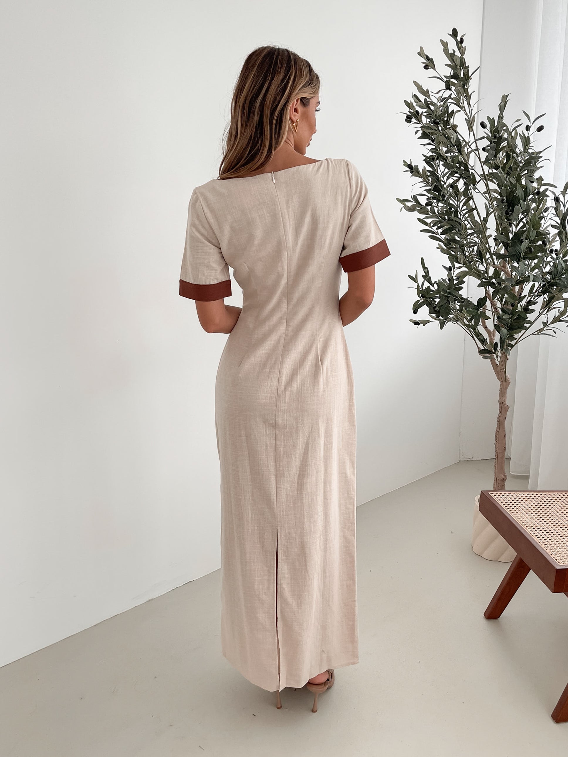 A woman with long, wavy blonde hair poses indoors in a minimalist room with white walls and a potted plant. She is wearing a light beige short-sleeve dress with brown trim details at the sleeves and waist, featuring a square neckline and a slim fit. She completes the look with pointed nude heels and a soft, confident expression.