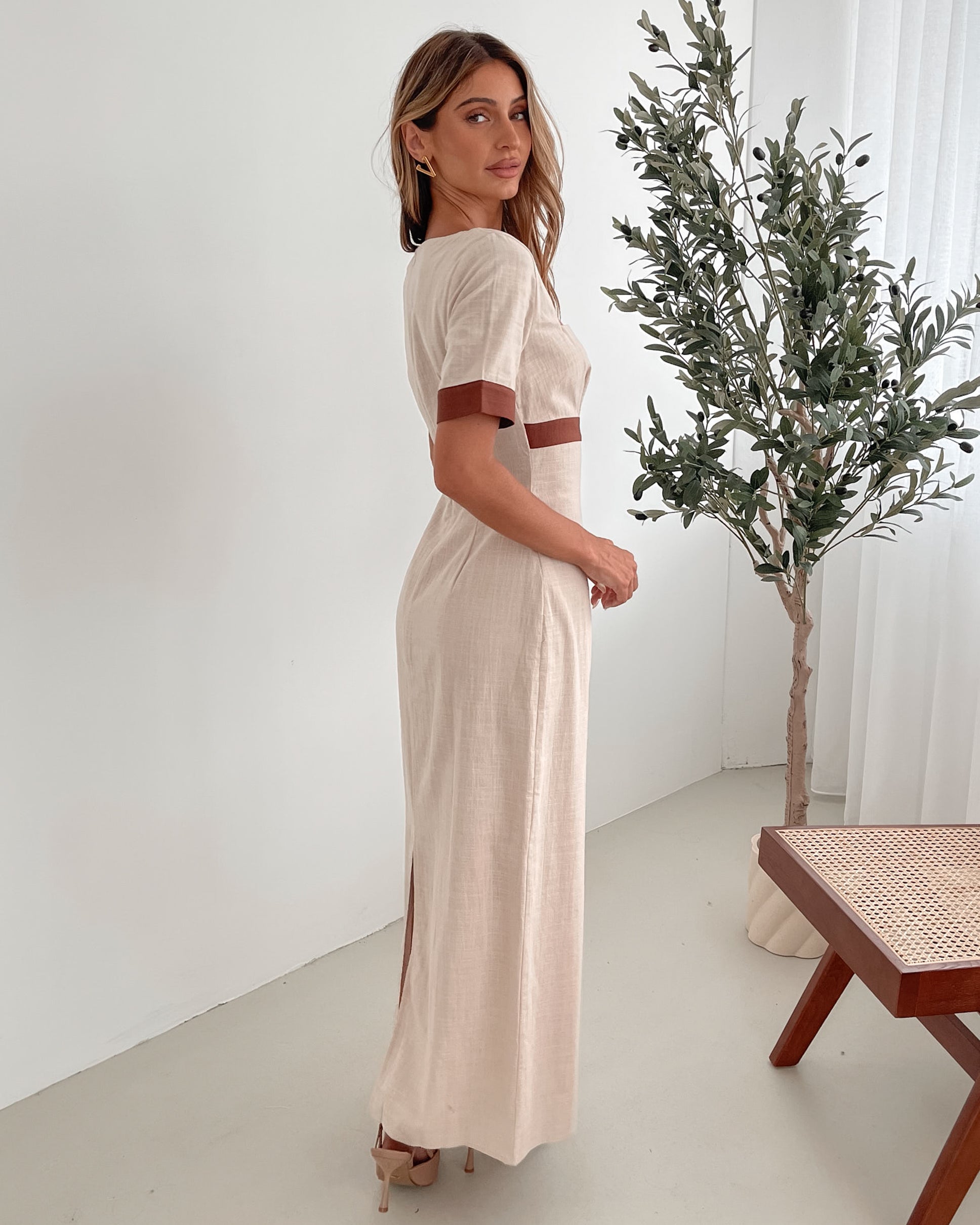 A woman with long, wavy blonde hair poses indoors in a minimalist room with white walls and a potted plant. She is wearing a light beige short-sleeve dress with brown trim details at the sleeves and waist, featuring a square neckline and a slim fit. She completes the look with pointed nude heels and a soft, confident expression.
