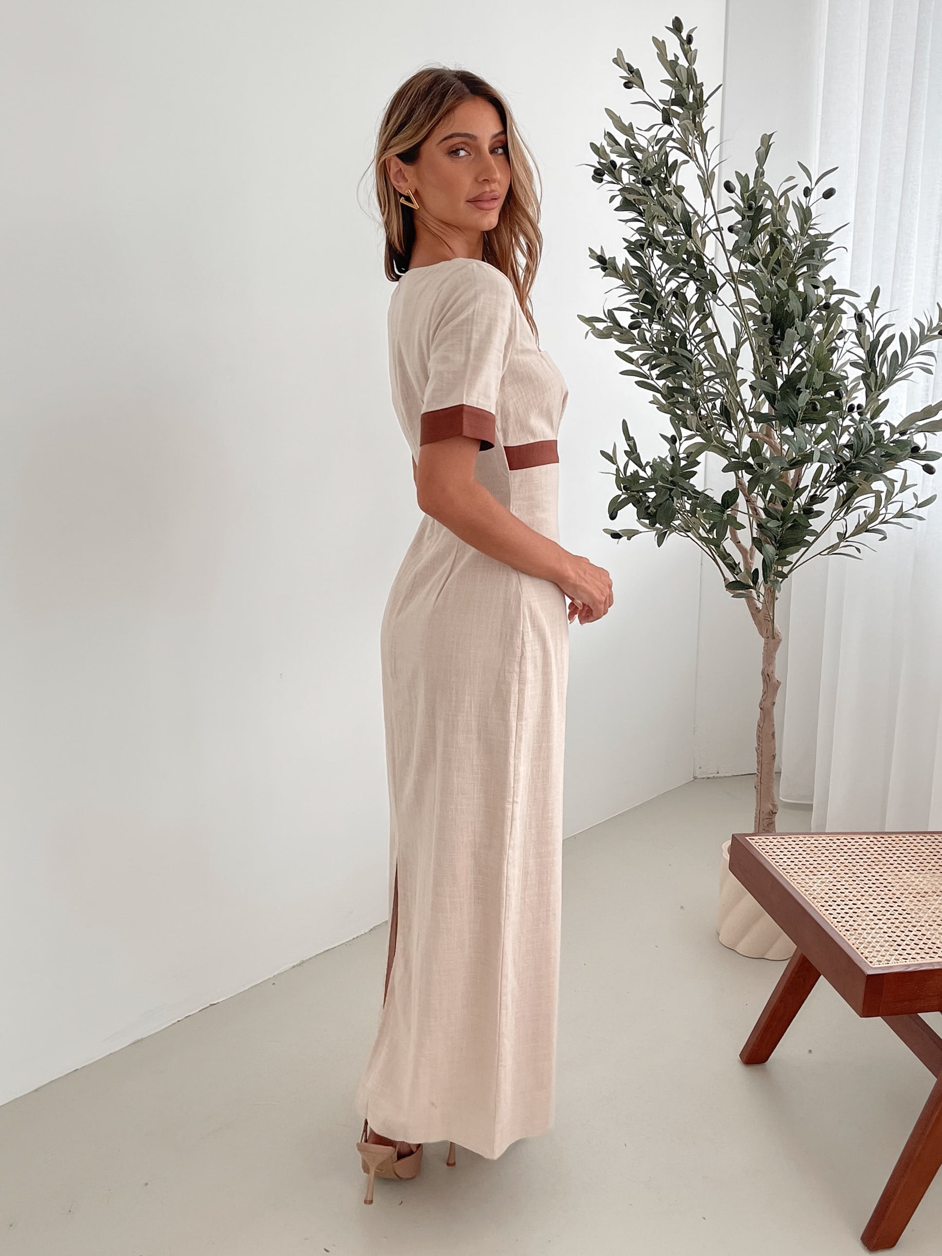 A woman with long, wavy blonde hair poses indoors in a minimalist room with white walls and a potted plant. She is wearing a light beige short-sleeve dress with brown trim details at the sleeves and waist, featuring a square neckline and a slim fit. She completes the look with pointed nude heels and a soft, confident expression.