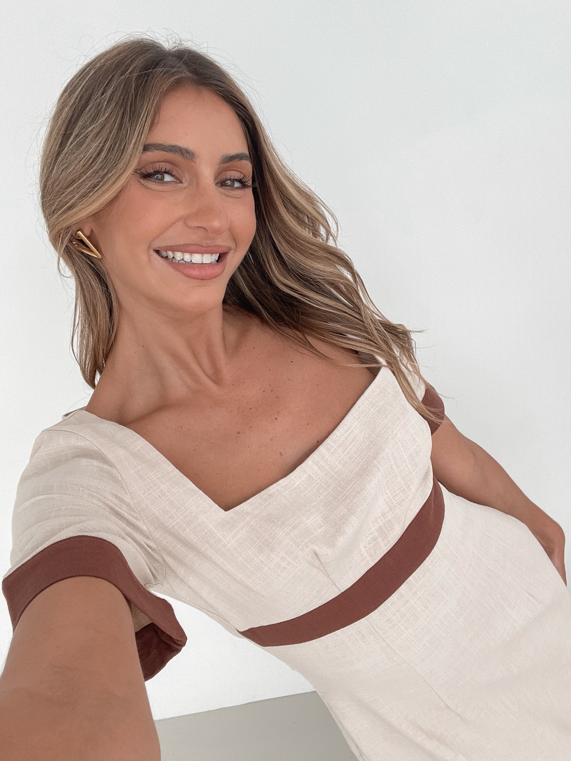 A woman with long, wavy blonde hair poses indoors in a minimalist room with white walls and a potted plant. She is wearing a light beige short-sleeve dress with brown trim details at the sleeves and waist, featuring a square neckline and a slim fit. She completes the look with pointed nude heels and a soft, confident expression.