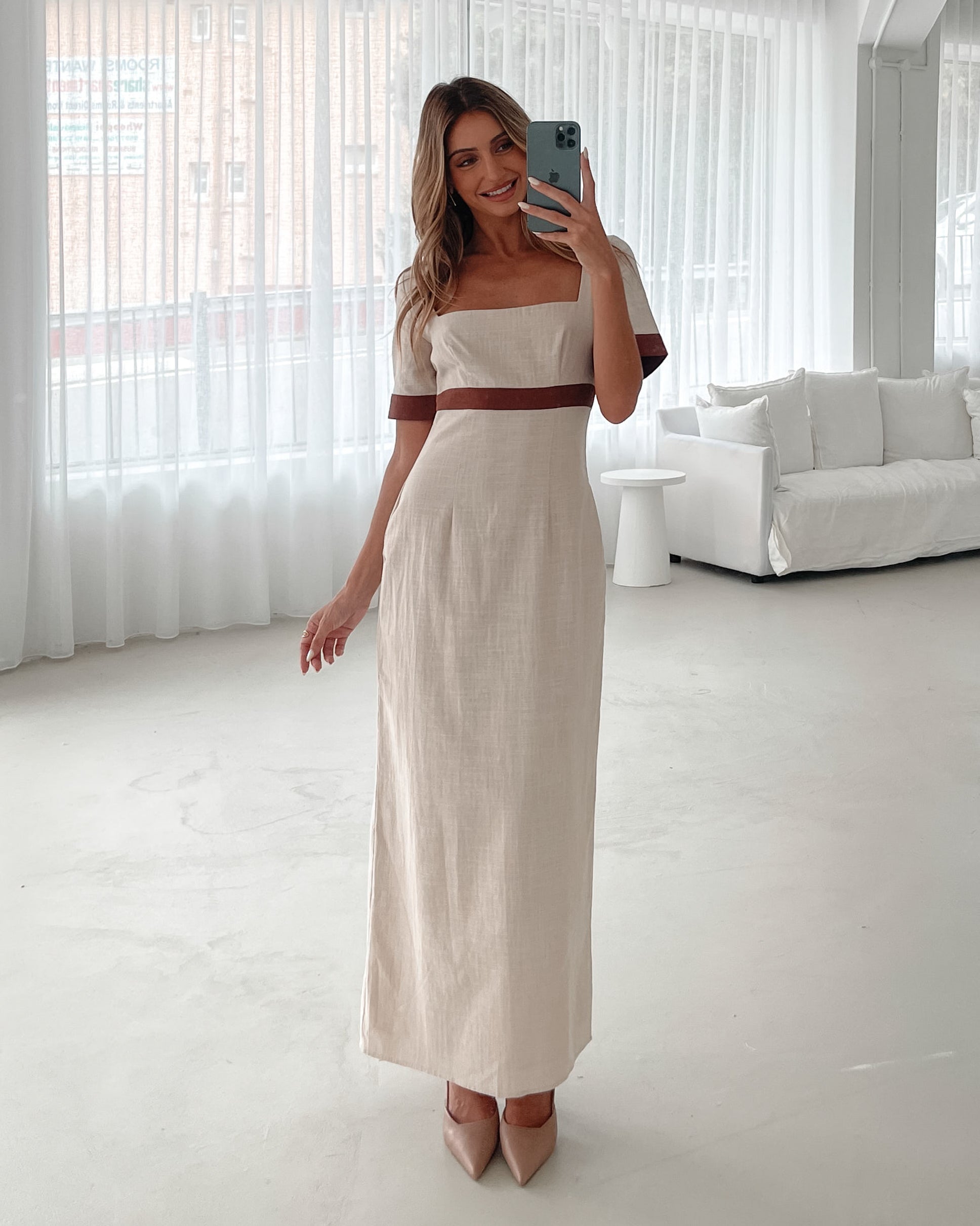 A woman with long, wavy blonde hair poses indoors in a minimalist room with white walls and a potted plant. She is wearing a light beige short-sleeve dress with brown trim details at the sleeves and waist, featuring a square neckline and a slim fit. She completes the look with pointed nude heels and a soft, confident expression.