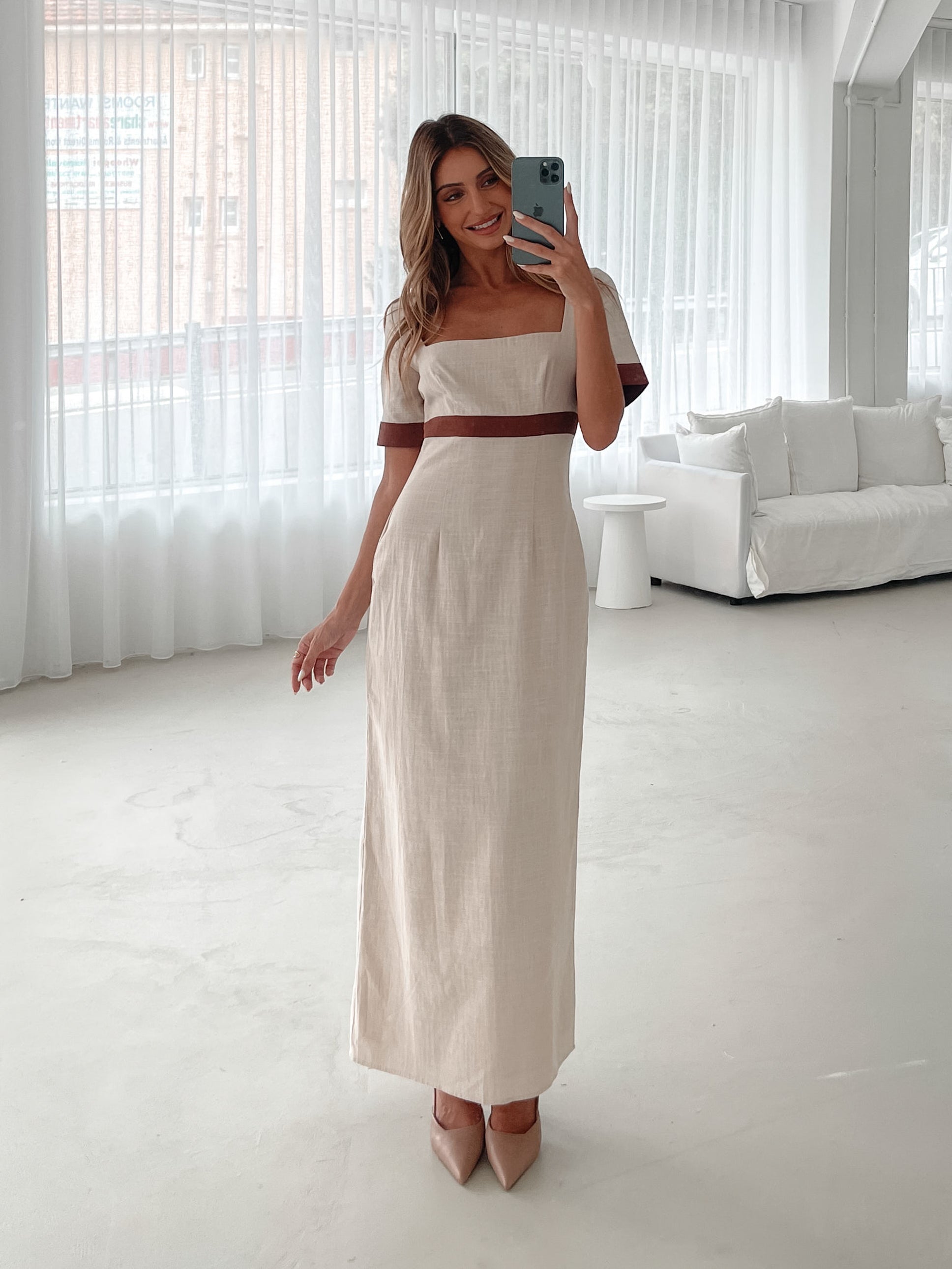 A woman with long, wavy blonde hair poses indoors in a minimalist room with white walls and a potted plant. She is wearing a light beige short-sleeve dress with brown trim details at the sleeves and waist, featuring a square neckline and a slim fit. She completes the look with pointed nude heels and a soft, confident expression.