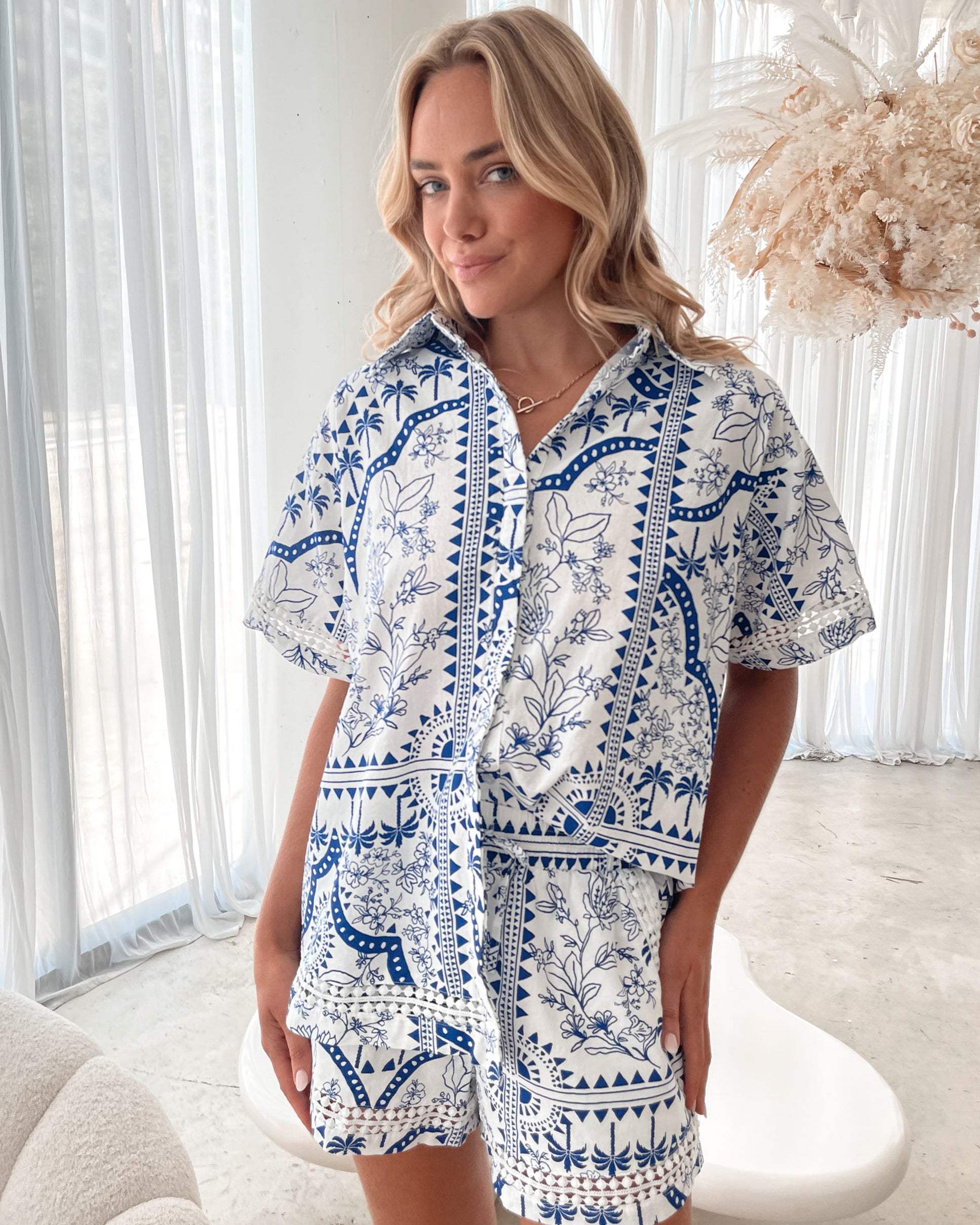 A woman with blonde, wavy hair stands in a bright, modern room with floor-to-ceiling sheer white curtains and neutral-toned decor. She is wearing a coordinated short-sleeve button-up shirt and matching shorts set featuring a blue and white floral and geometric print. She has a relaxed expression with one hand in her pocket, exuding a stylish and effortless vibe.
