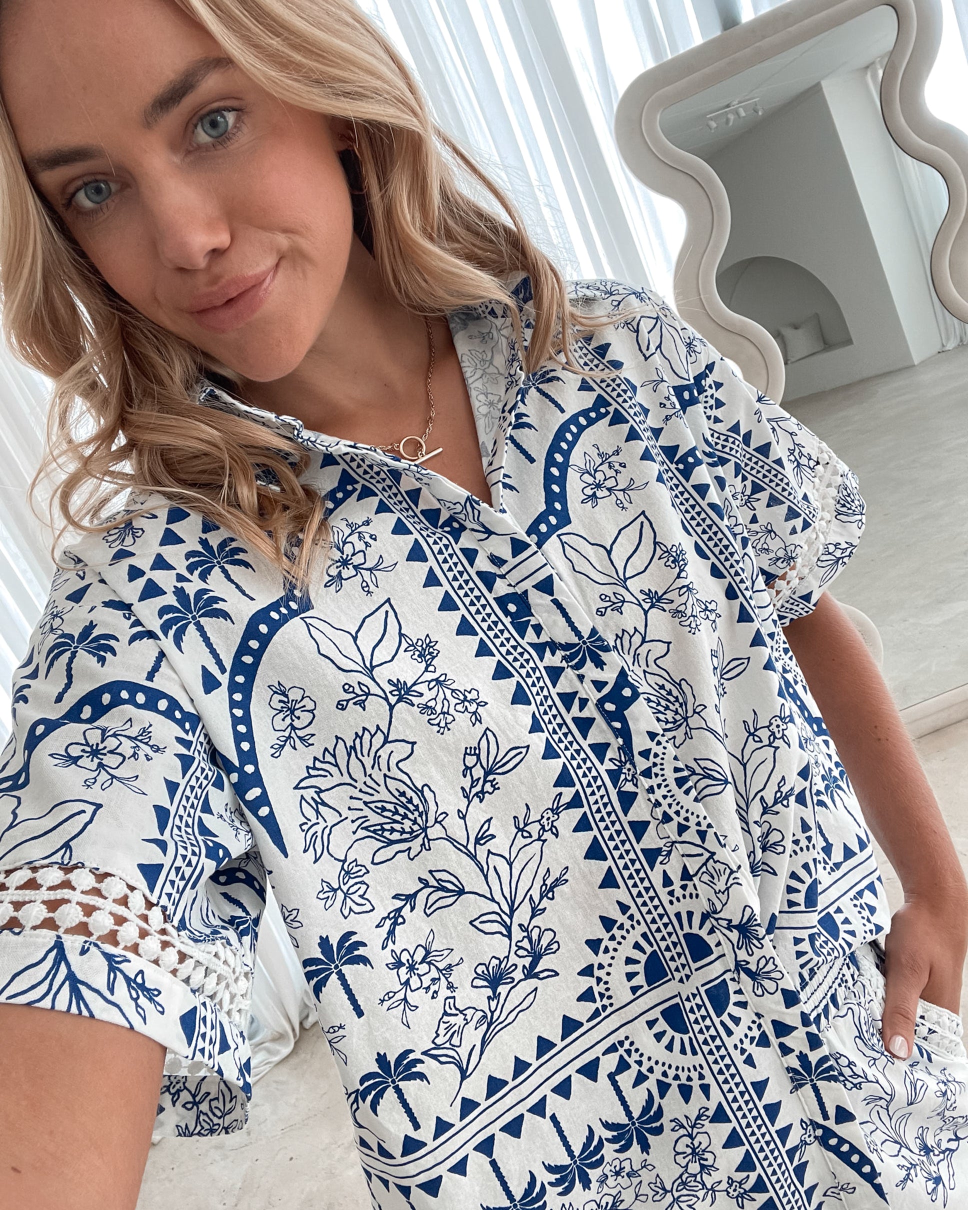 A woman with blonde, wavy hair stands in a bright, modern room with floor-to-ceiling sheer white curtains and neutral-toned decor. She is wearing a coordinated short-sleeve button-up shirt and matching shorts set featuring a blue and white floral and geometric print. She has a relaxed expression with one hand in her pocket, exuding a stylish and effortless vibe.