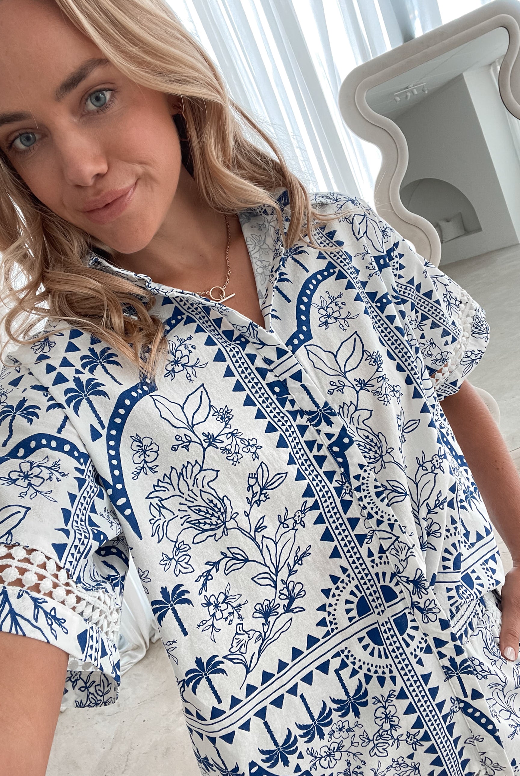 A woman with blonde, wavy hair stands in a bright, modern room with floor-to-ceiling sheer white curtains and neutral-toned decor. She is wearing a coordinated short-sleeve button-up shirt and matching shorts set featuring a blue and white floral and geometric print. She has a relaxed expression with one hand in her pocket, exuding a stylish and effortless vibe.