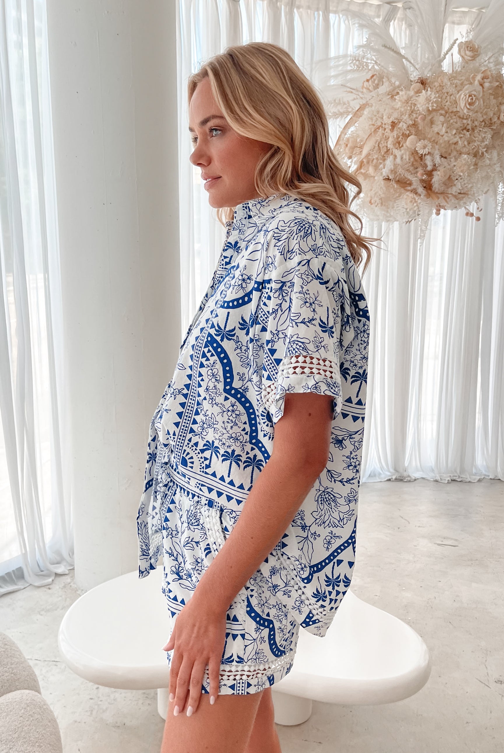 A woman with blonde, wavy hair stands in a bright, modern room with floor-to-ceiling sheer white curtains and neutral-toned decor. She is wearing a coordinated short-sleeve button-up shirt and matching shorts set featuring a blue and white floral and geometric print. She has a relaxed expression with one hand in her pocket, exuding a stylish and effortless vibe.