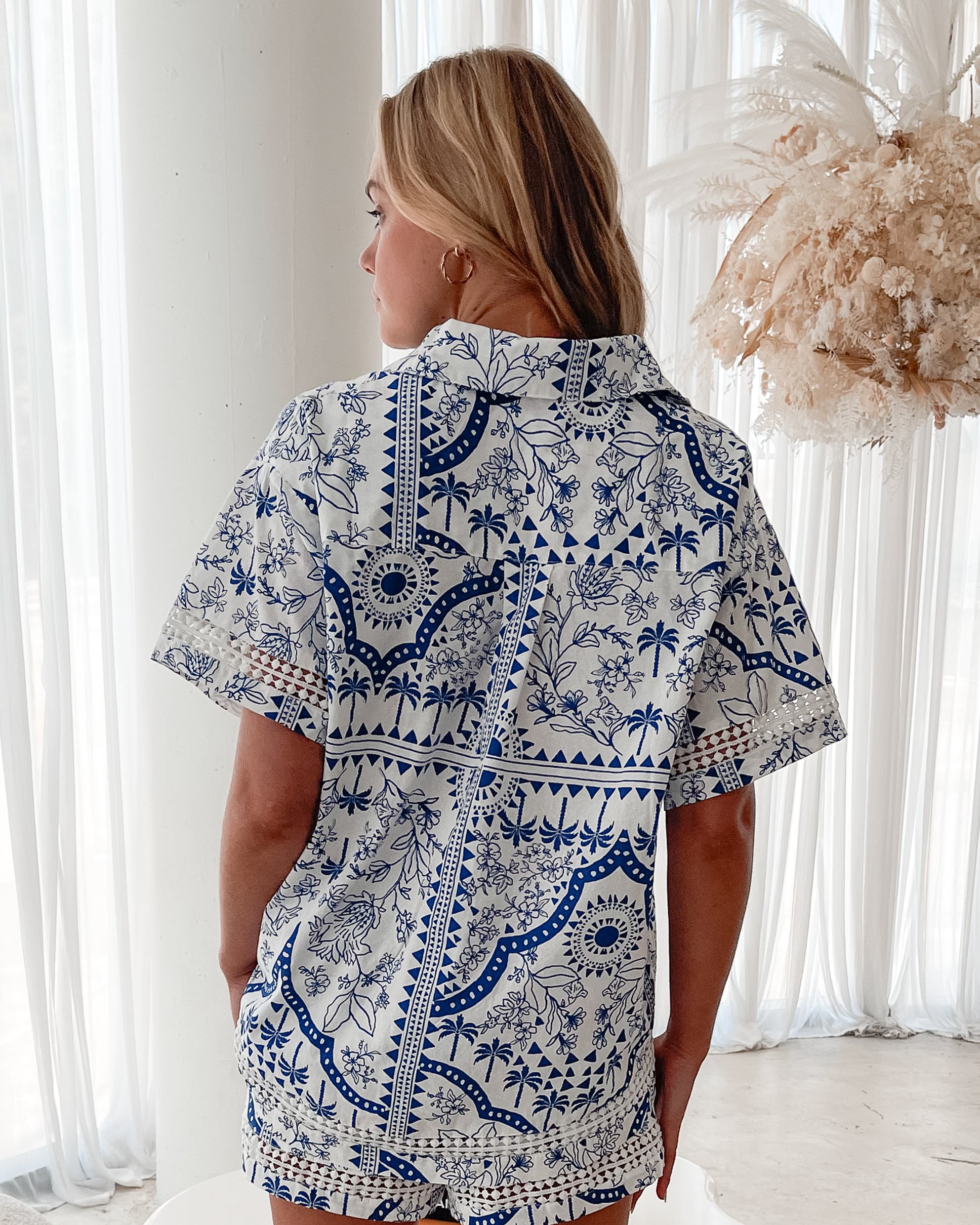 A woman with blonde, wavy hair stands in a bright, modern room with floor-to-ceiling sheer white curtains and neutral-toned decor. She is wearing a coordinated short-sleeve button-up shirt and matching shorts set featuring a blue and white floral and geometric print. She has a relaxed expression with one hand in her pocket, exuding a stylish and effortless vibe.